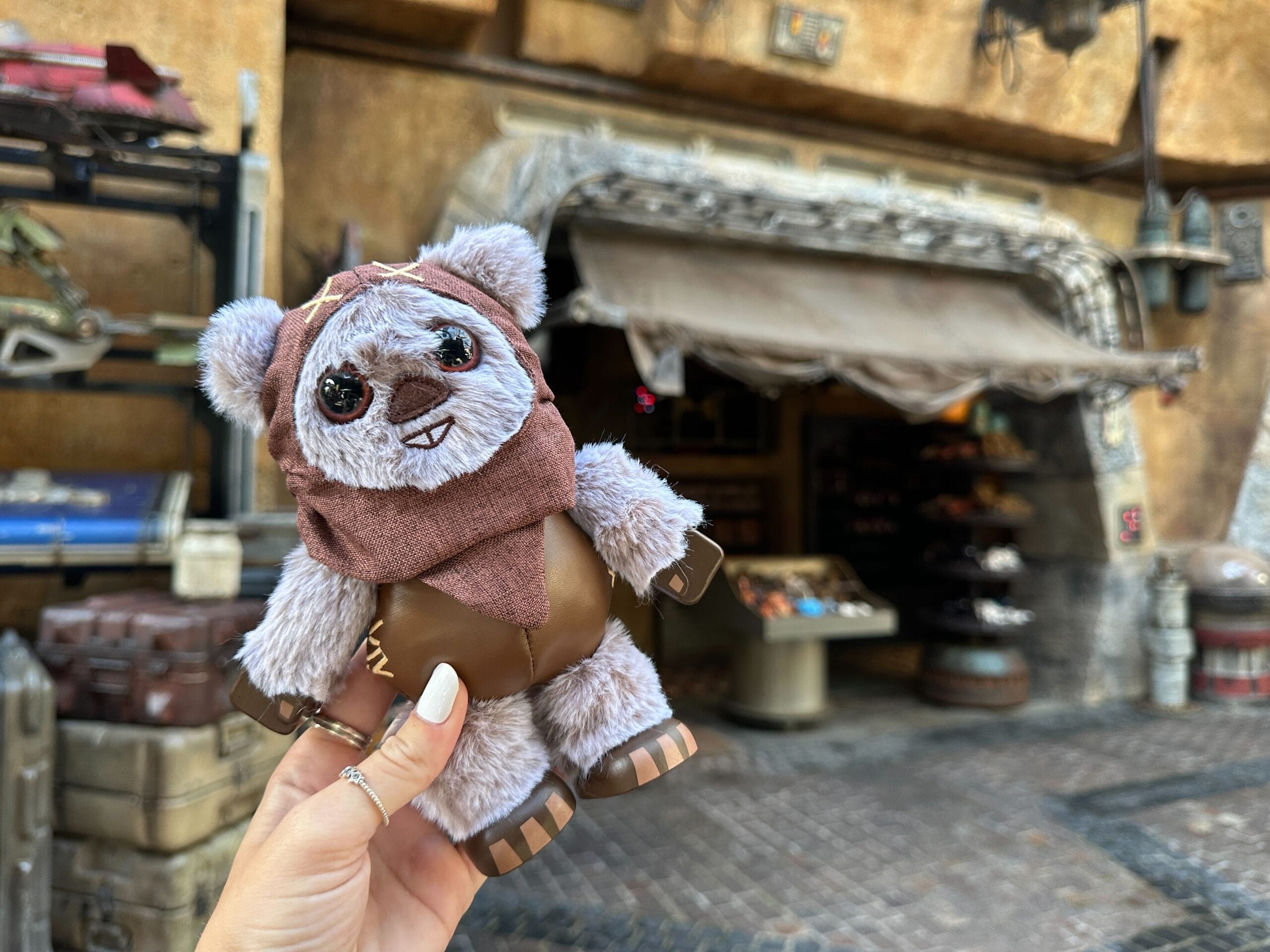 A hand holds an Ewok Plush in front of a rustic shop at Star Wars Galaxy’s Edge in Walt Disney World.