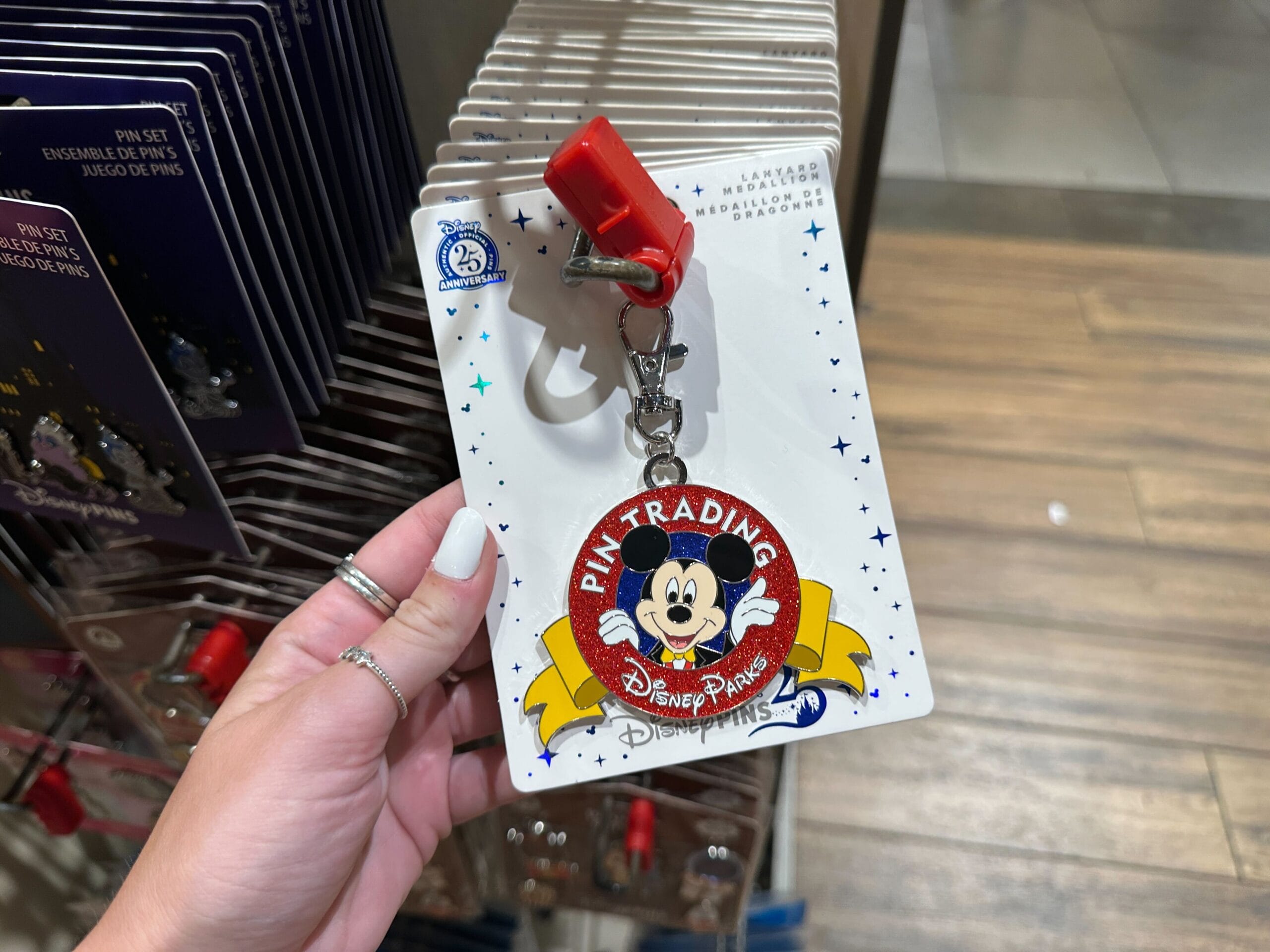 A hand holds a Disney Pin Trading keychain featuring Mickey Mouse and a red clip in a store display.