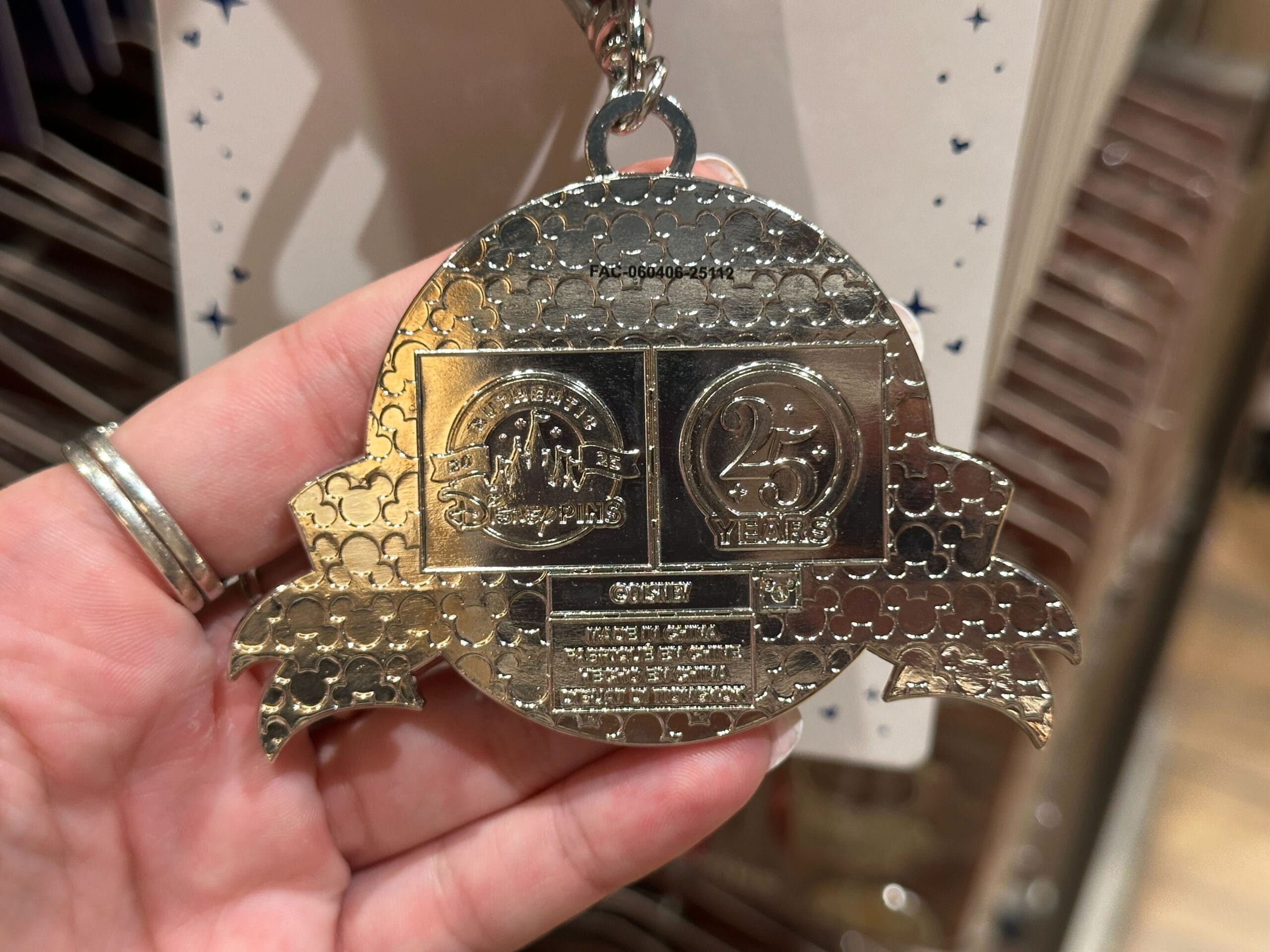 A hand holding a silver 25th anniversary keychain with embossed logos and decorative patterns.