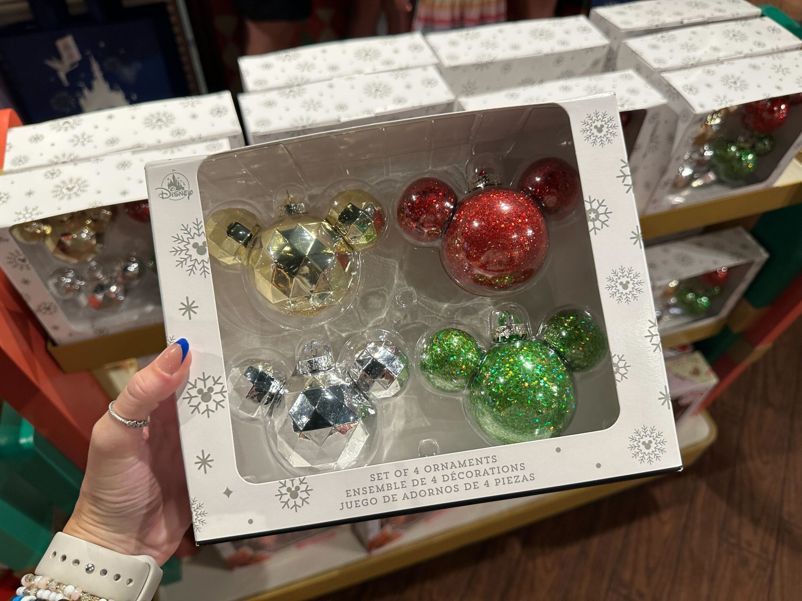 A hand holds a box of four Mickey Mouse-shaped Christmas ornaments in red, gold, green, and silver.