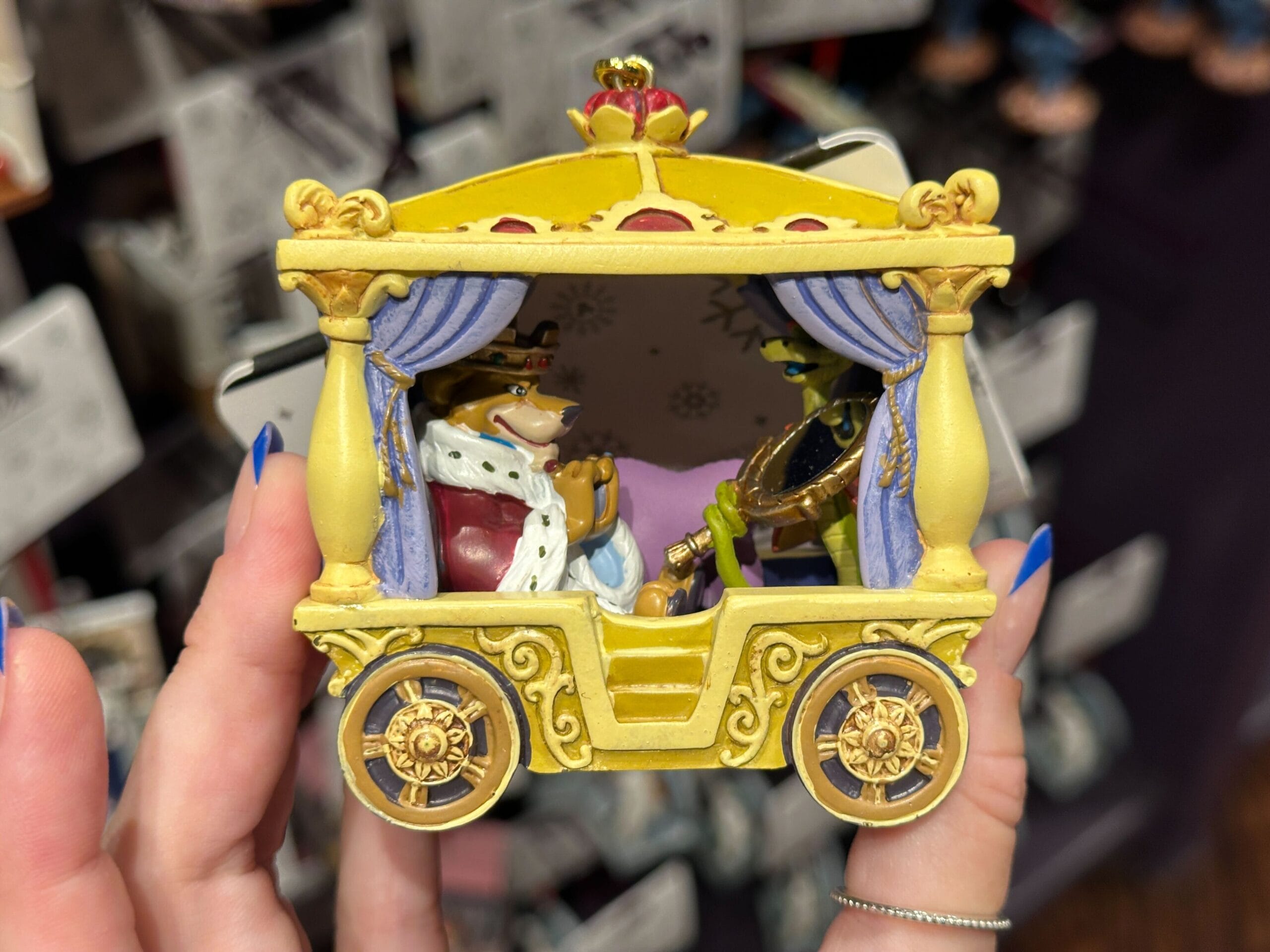A hand holds a yellow carriage ornament featuring two animated characters inside, one wearing a crown and robe.