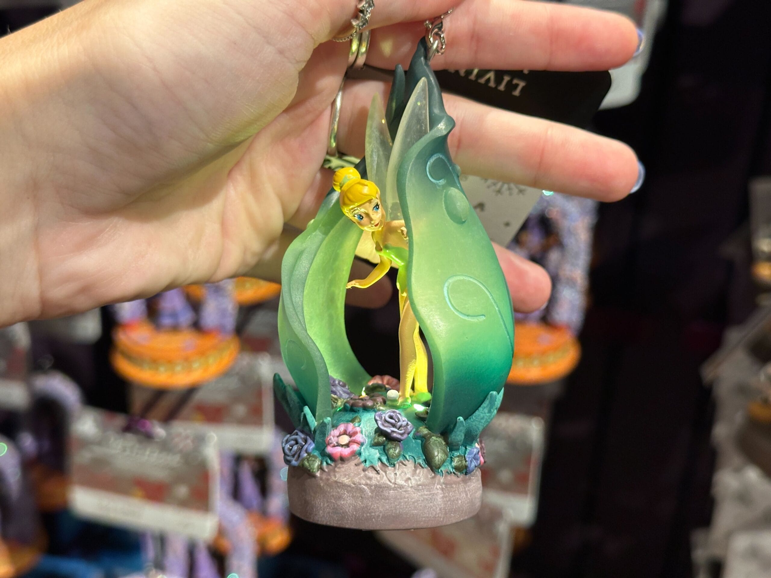 A hand holds a Tinker Bell figurine surrounded by green leaves and colorful flowers.