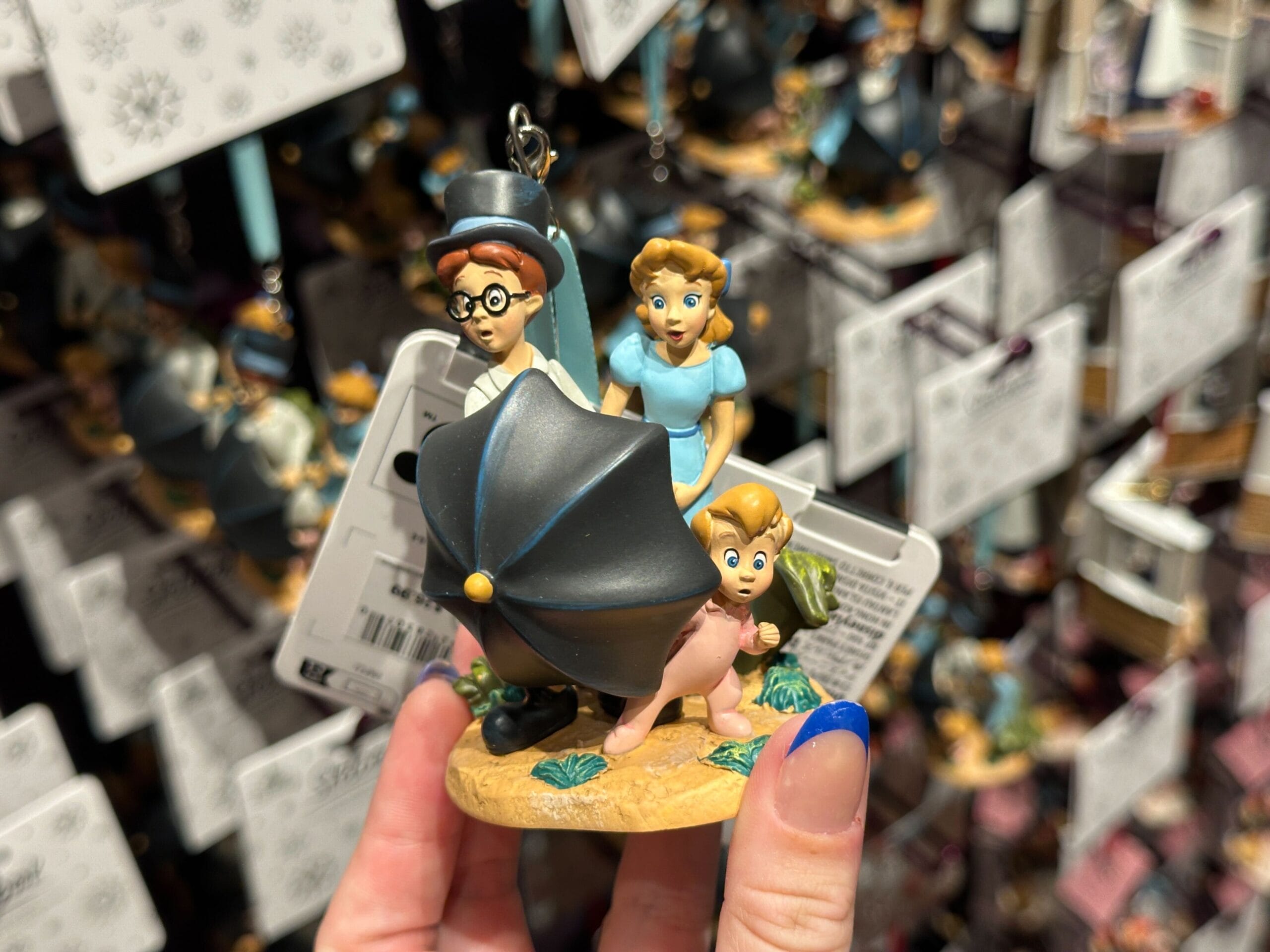 A hand holds a Peter Pan-themed ornament with three characters and an umbrella, against a background of similar ornaments.