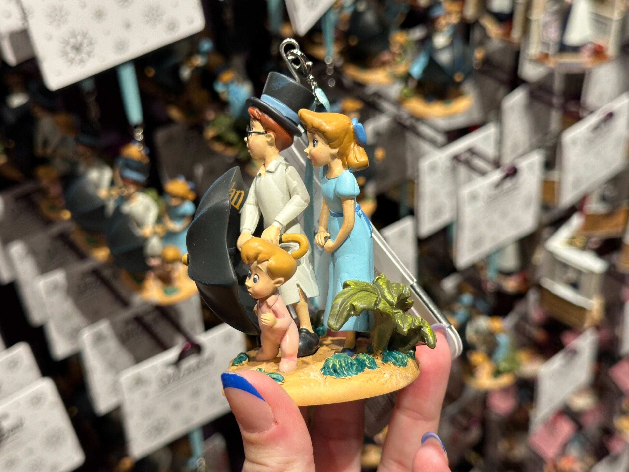 Hand holding a Peter Pan-themed ornament featuring Wendy, John, and Michael standing together, with ornaments in the background.