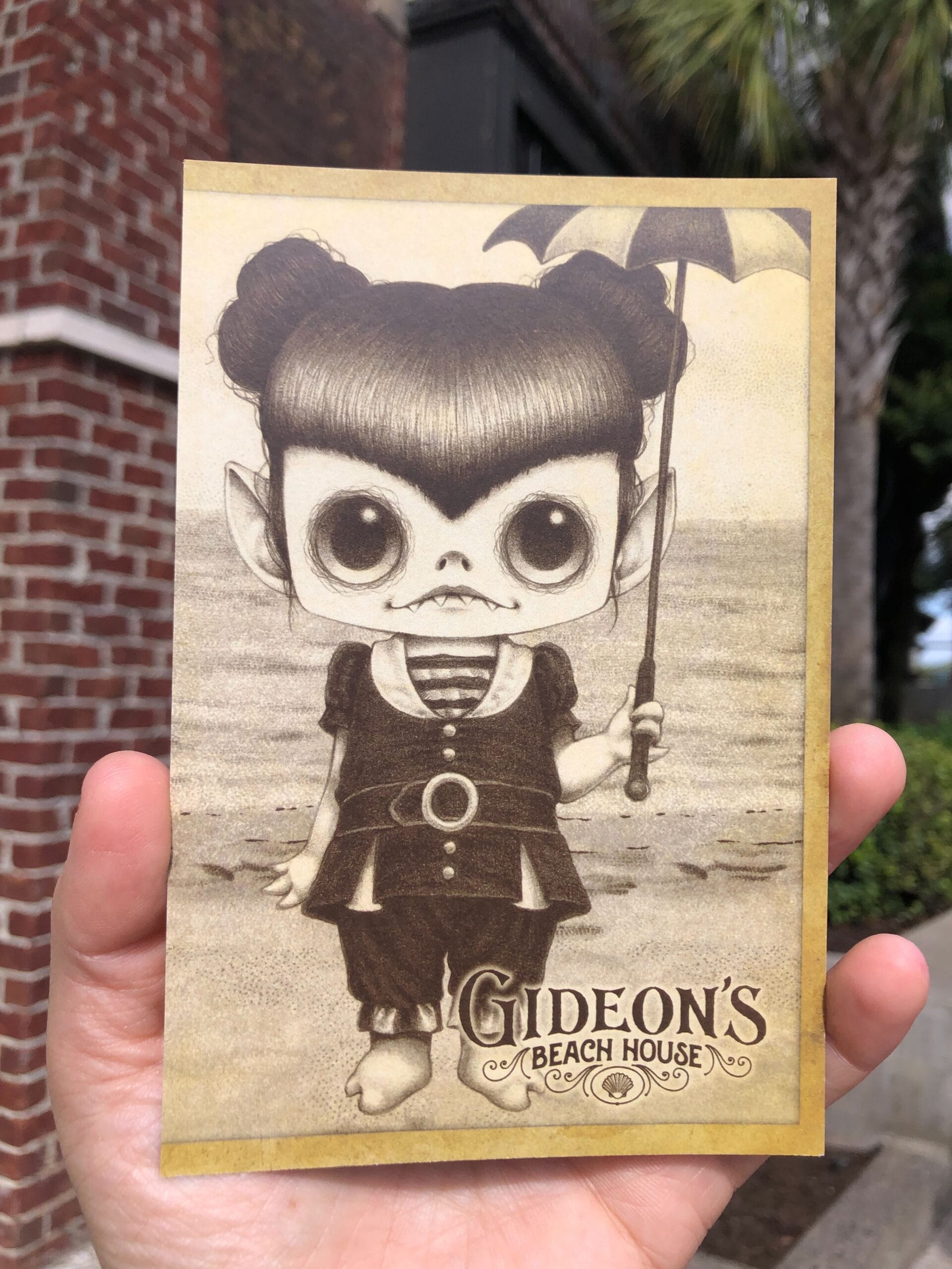A hand holds a postcard of a big-eyed cartoon girl with fangs, "Gideon's Bakehouse" and July 2025 written below.