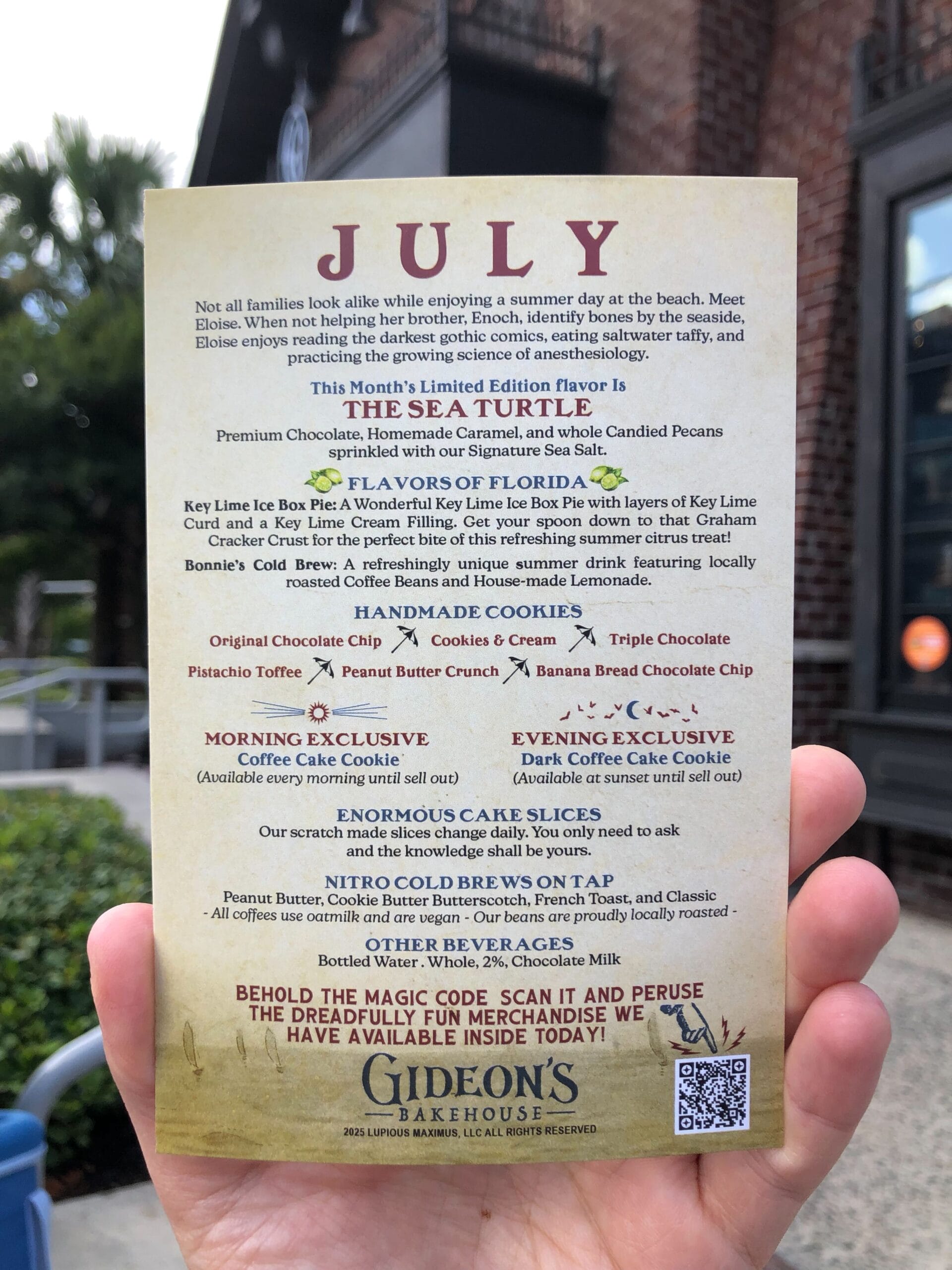 A hand holds a Gideon's Bakehouse July 2025 menu, featuring the Sea Turtle Cookie, in front of a brick building and trees.
