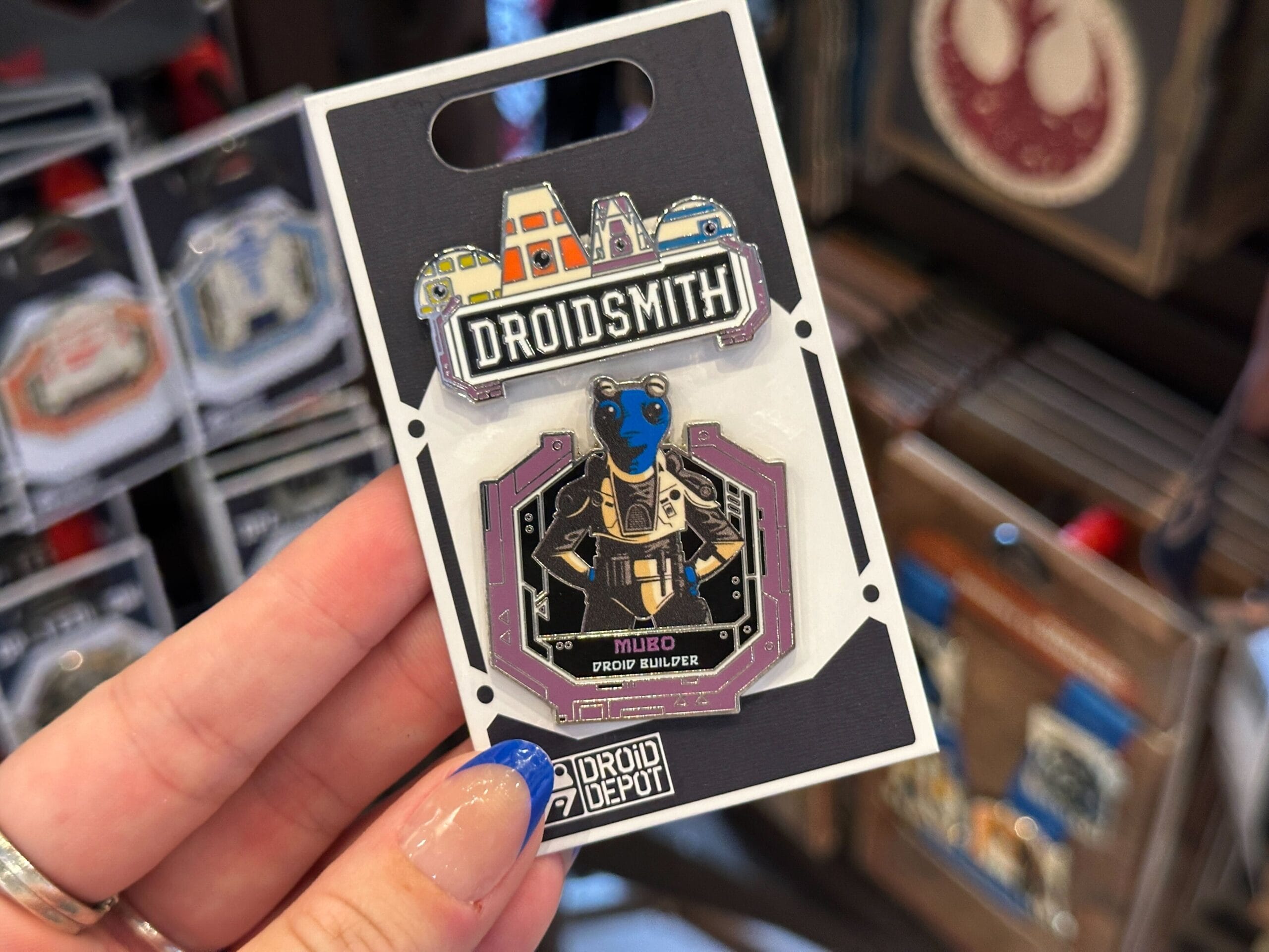 A hand holds a Star Wars Droid Depot "Mubo Droid Builder" pin on display among other pins in-store.