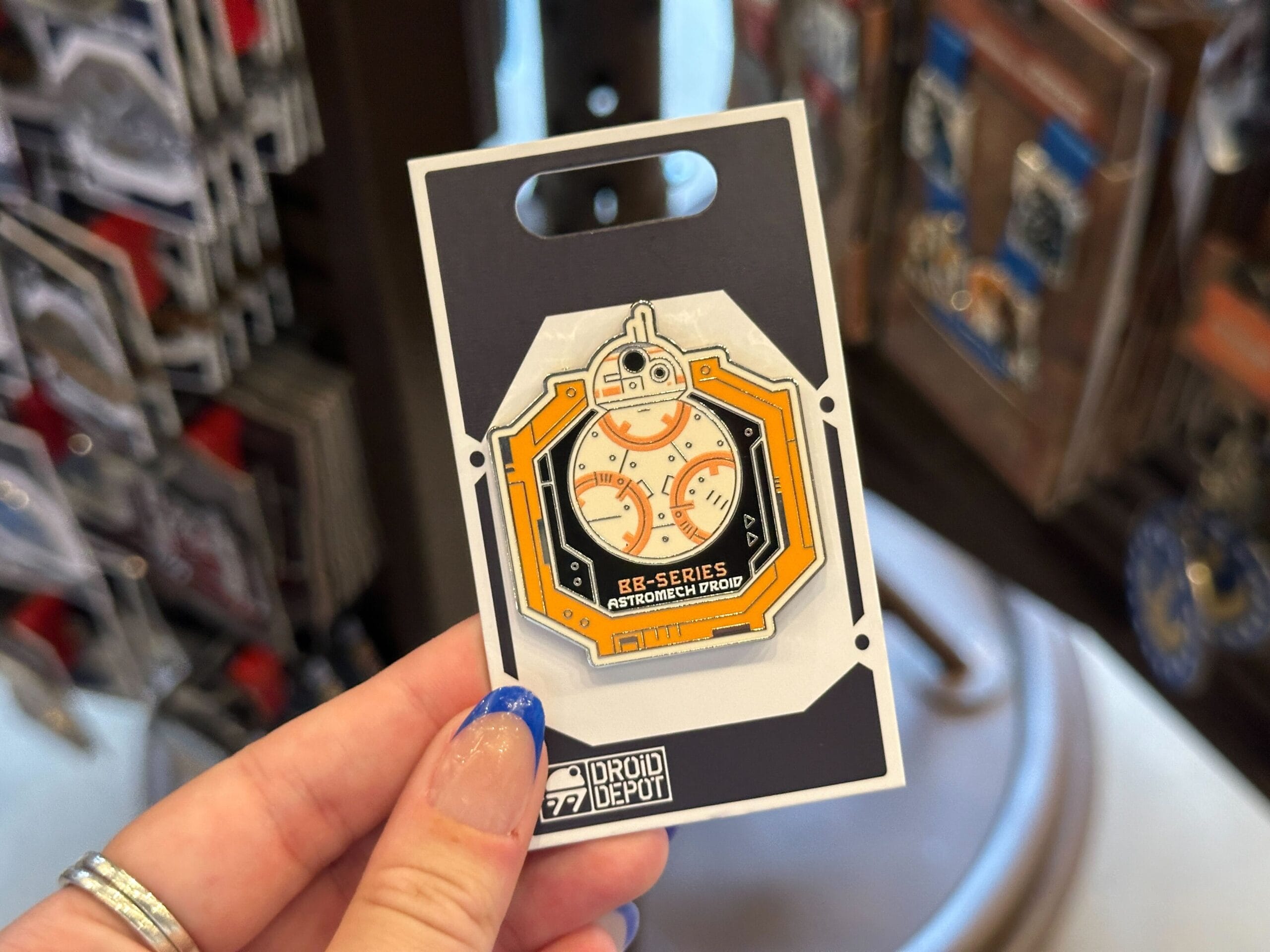 A hand holds a BB-series Droid Depot pin enamel pin, displayed on a themed Droid Depot Pins backing card.
