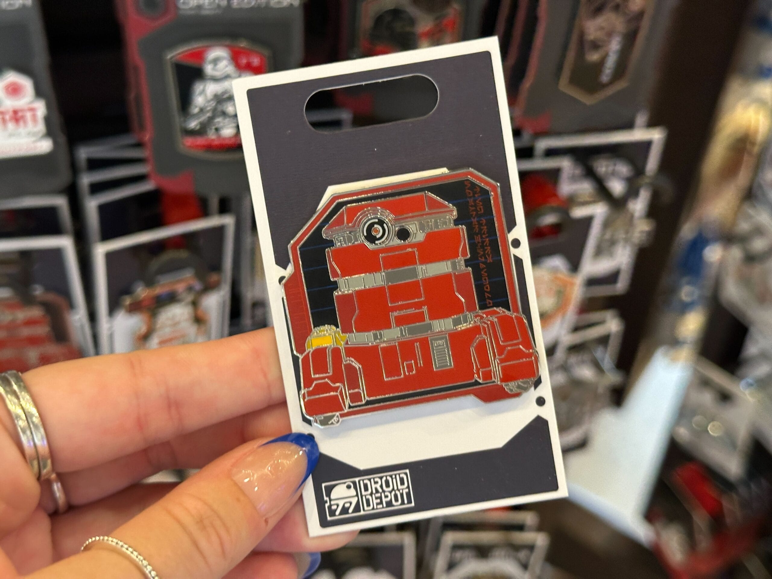 A hand holds a red Droid Depot pin on a Droid Depot card display, perfect for any Star Wars pins collection.