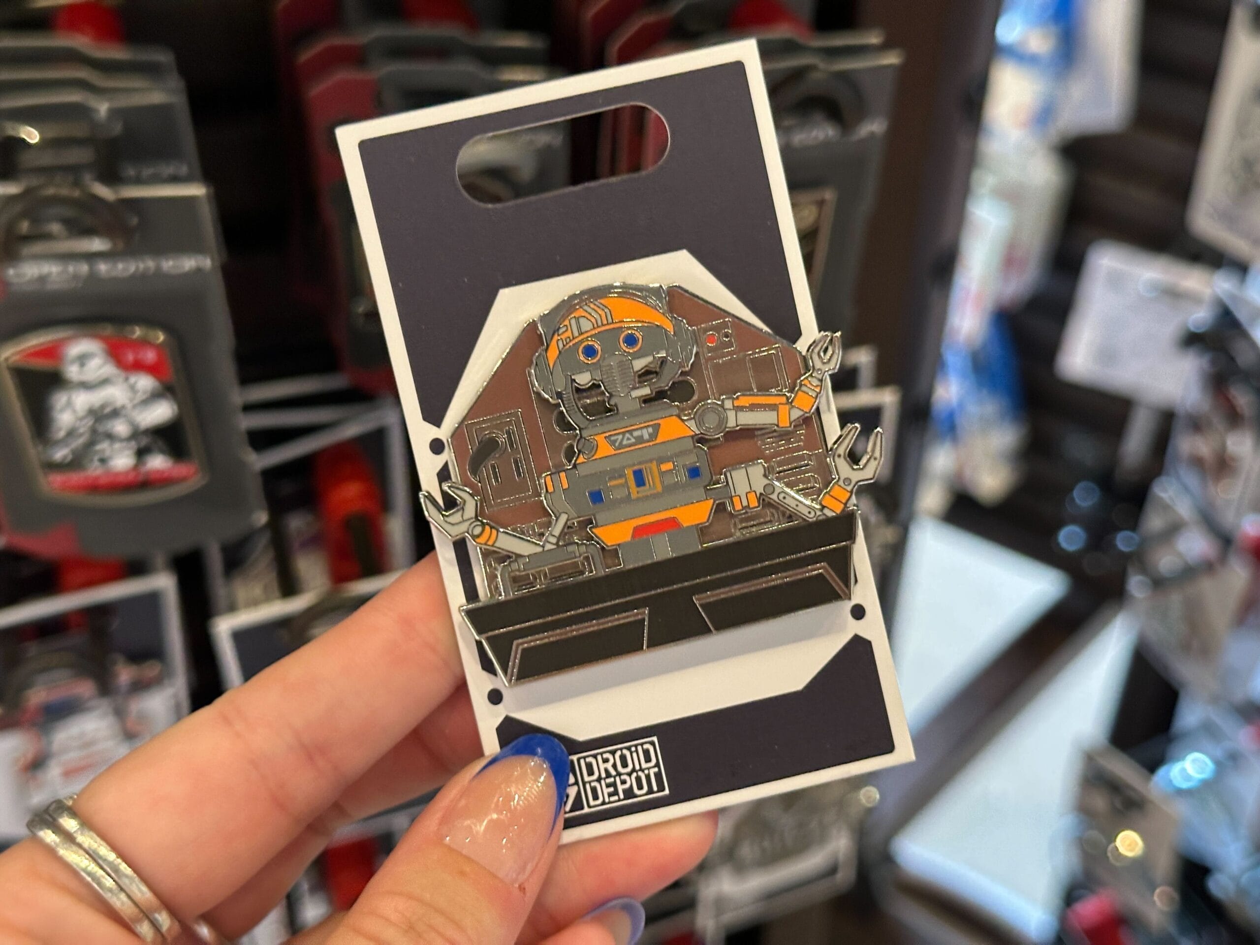 A hand holds a Star Wars Droid Depot pin featuring an orange and blue droid, with other pins displayed in the background.
