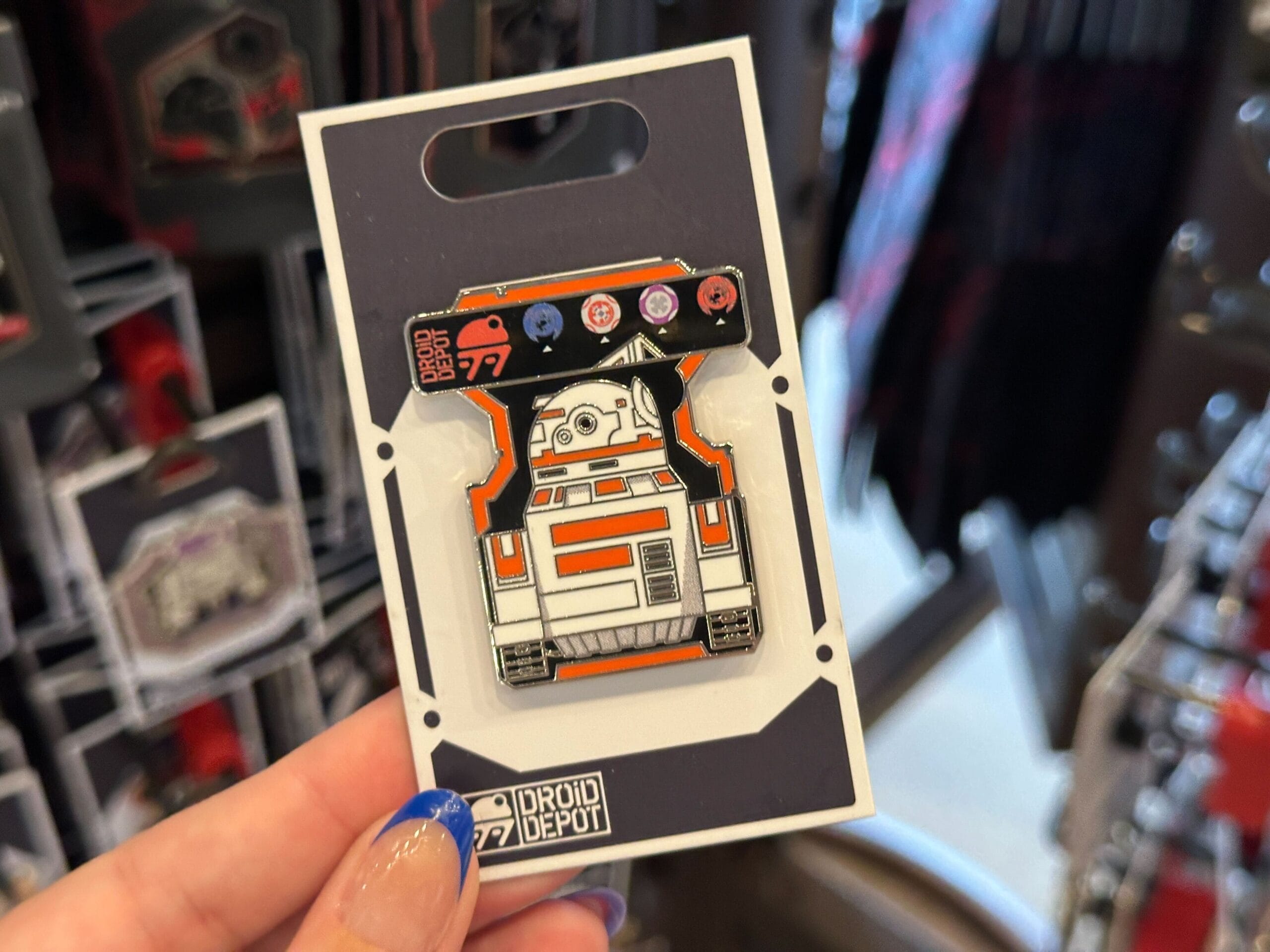 A hand holds a Droid Depot pin featuring an orange and white Star Wars droid, displayed on a black card.