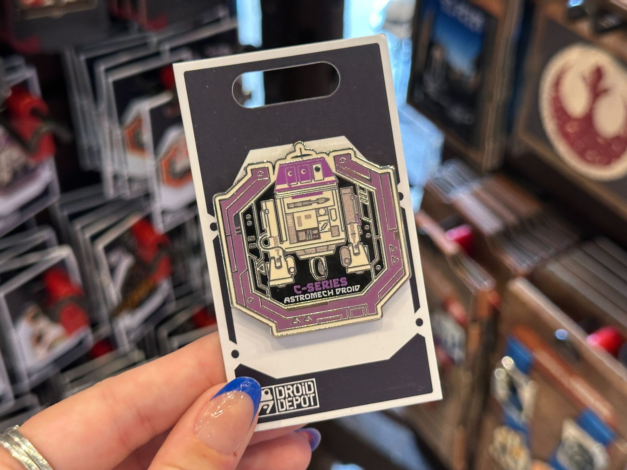 A hand holds a purple and silver Star Wars C-Series Astromech Droid pin from Droid Depot in a store display.
