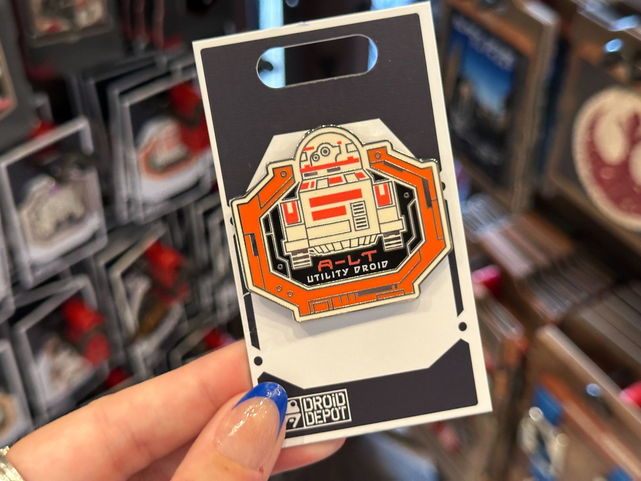 Close-up of a hand holding a Droid Depot Pin at a store display, with other Star Wars pins blurred in the background.