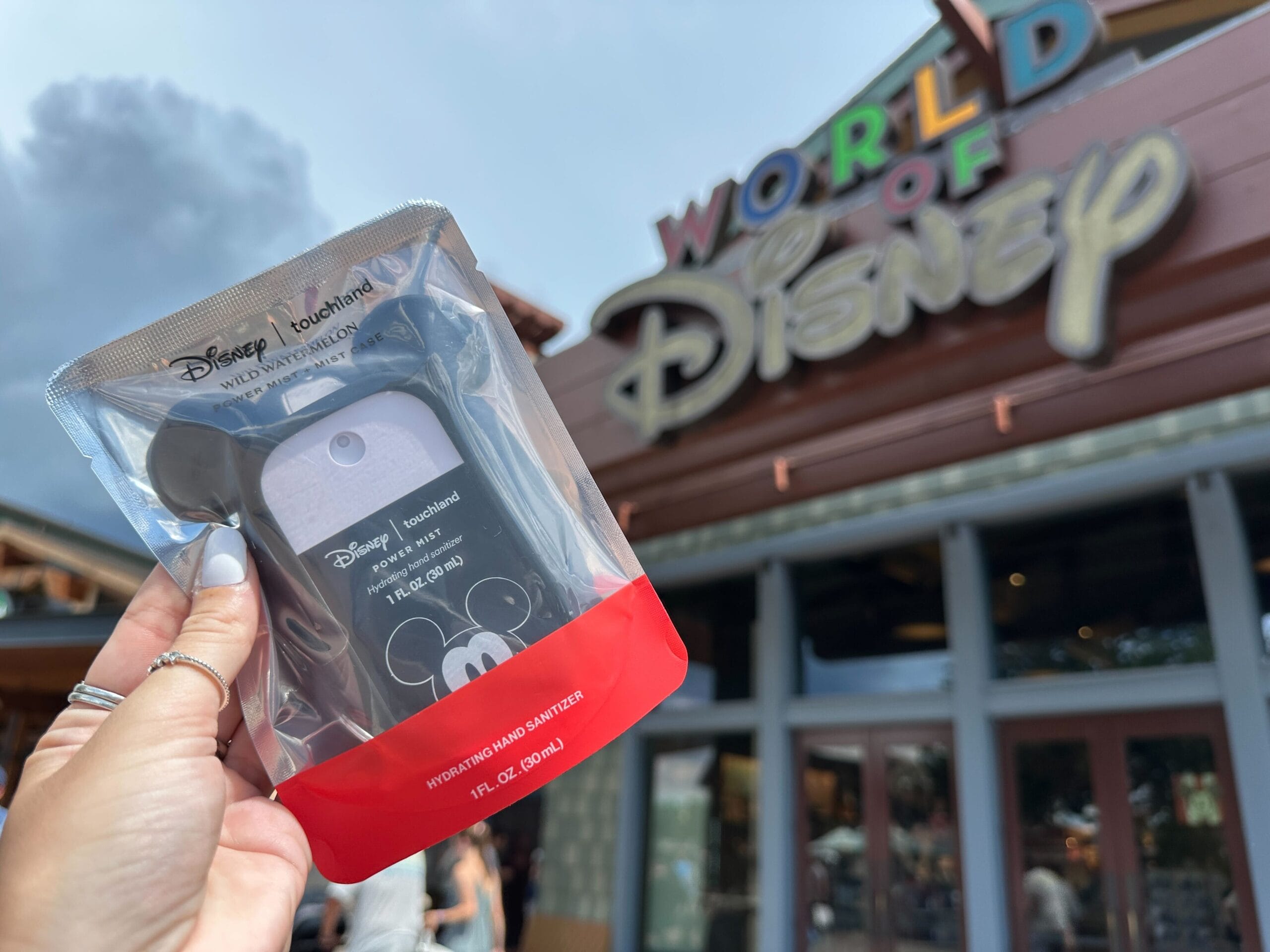 A hand holds Disney hydrating hand sanitizer in front of the World of Disney store entrance.