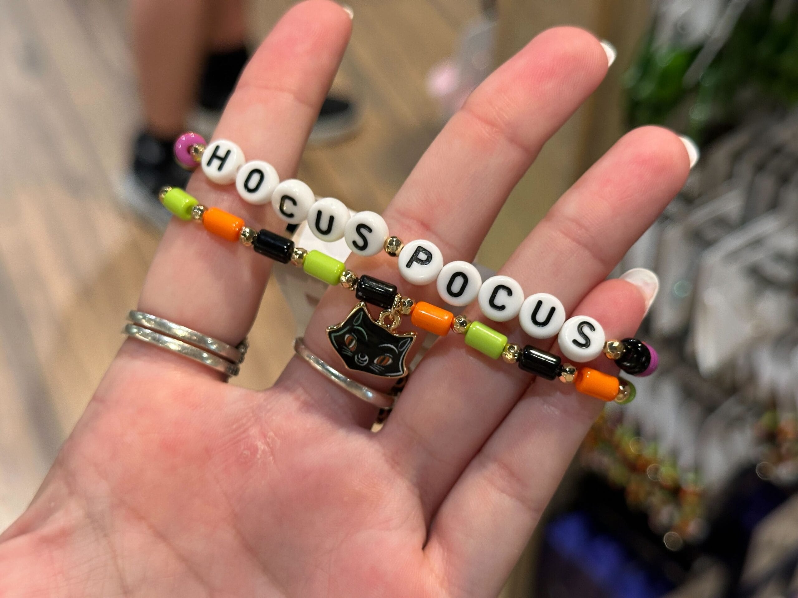 A hand holds a beaded bracelet that spells "HOCUS POCUS" with a black cat charm in the center.