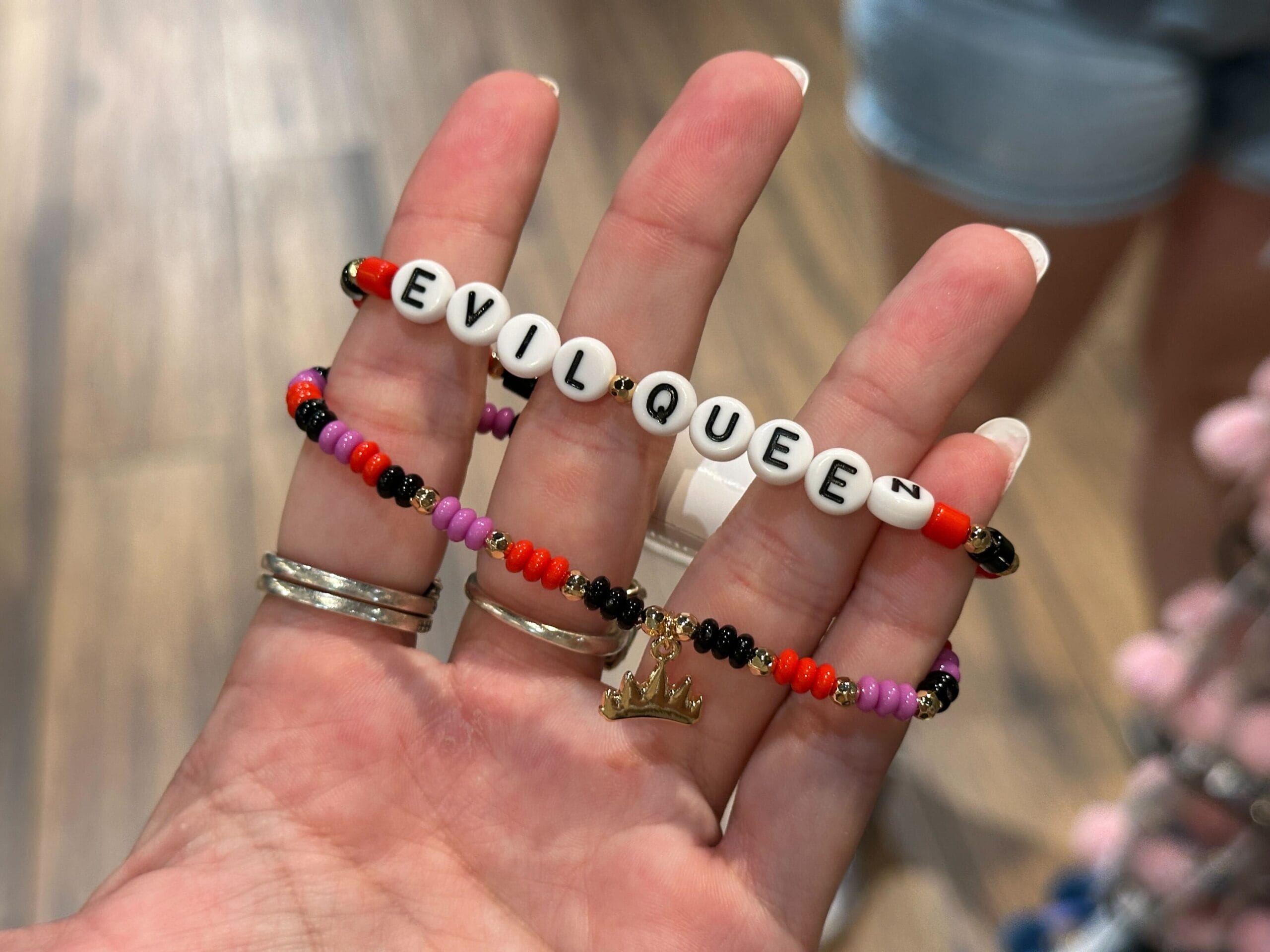 A hand holds two beaded bracelets, one spells “EVIL QUEEN” and the other has a gold crown charm.
