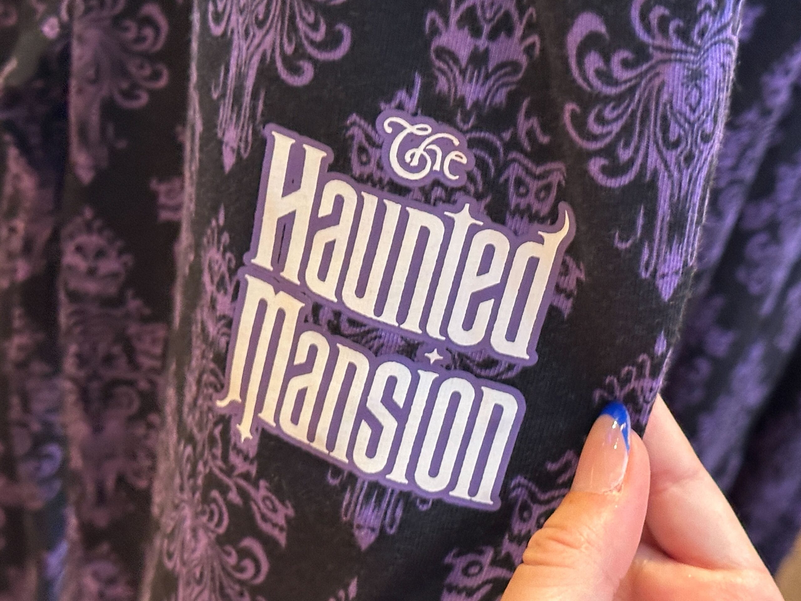 A hand holds Magic Kingdom Haunted Mansion fabric with the logo in white over a purple gothic pattern.