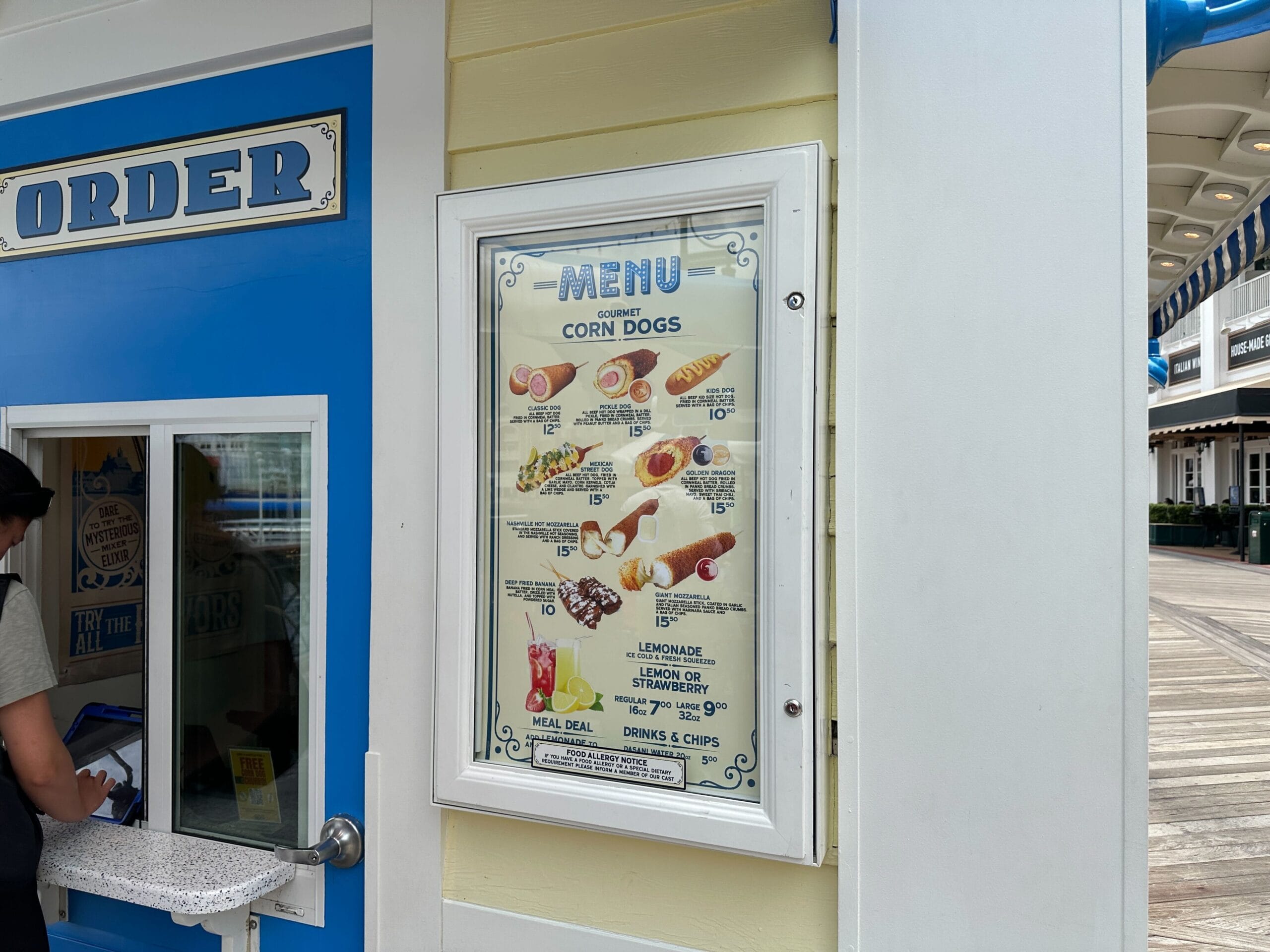A menu board for Blue Ribbon Corn Dog, featuring Nashville Hot options, is displayed by a blue and yellow walk-up window.