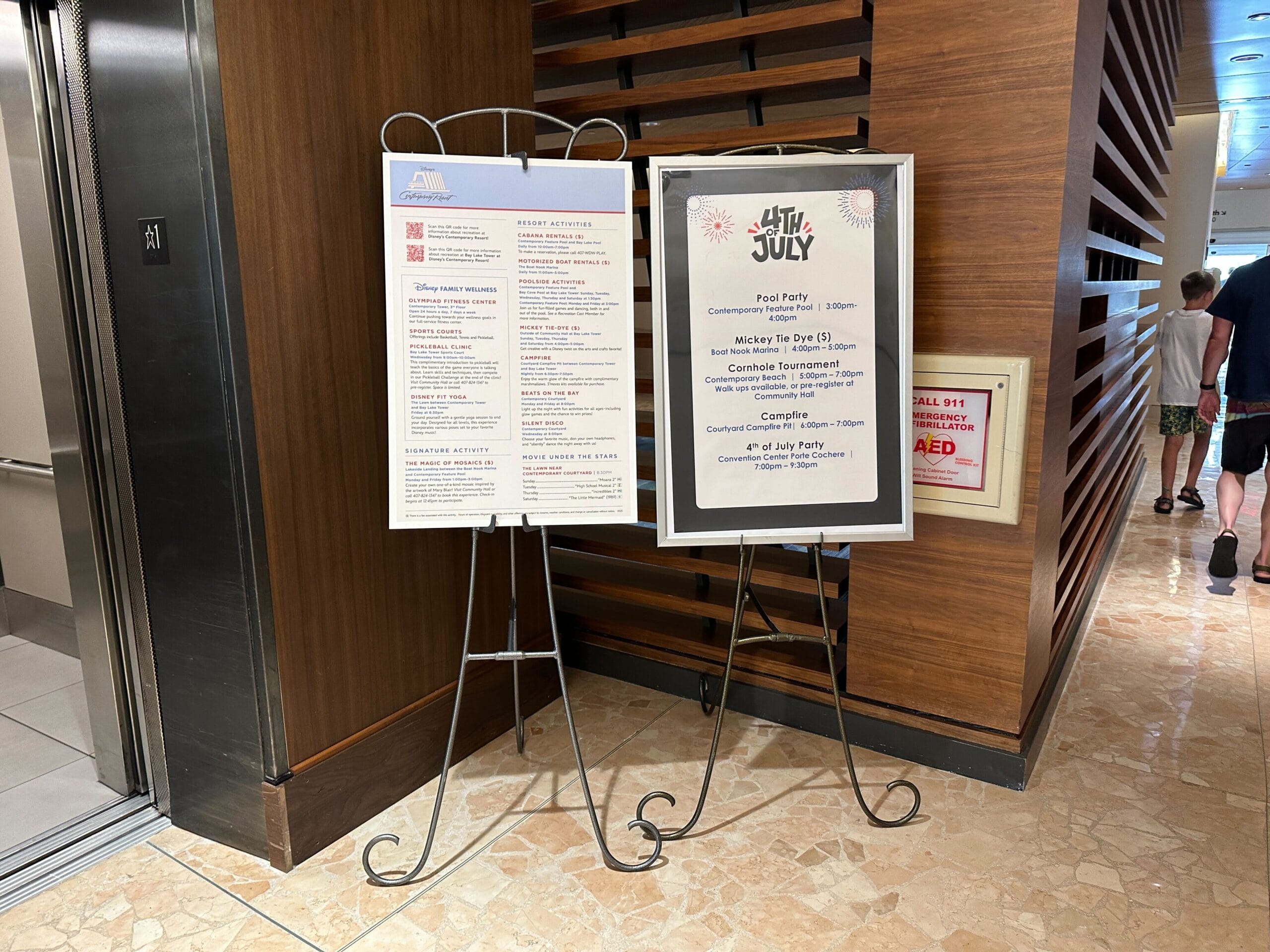 Two easels display Fourth of July 2025 event posters in a Walt Disney World Resort Hotels hallway near the elevator and AED.