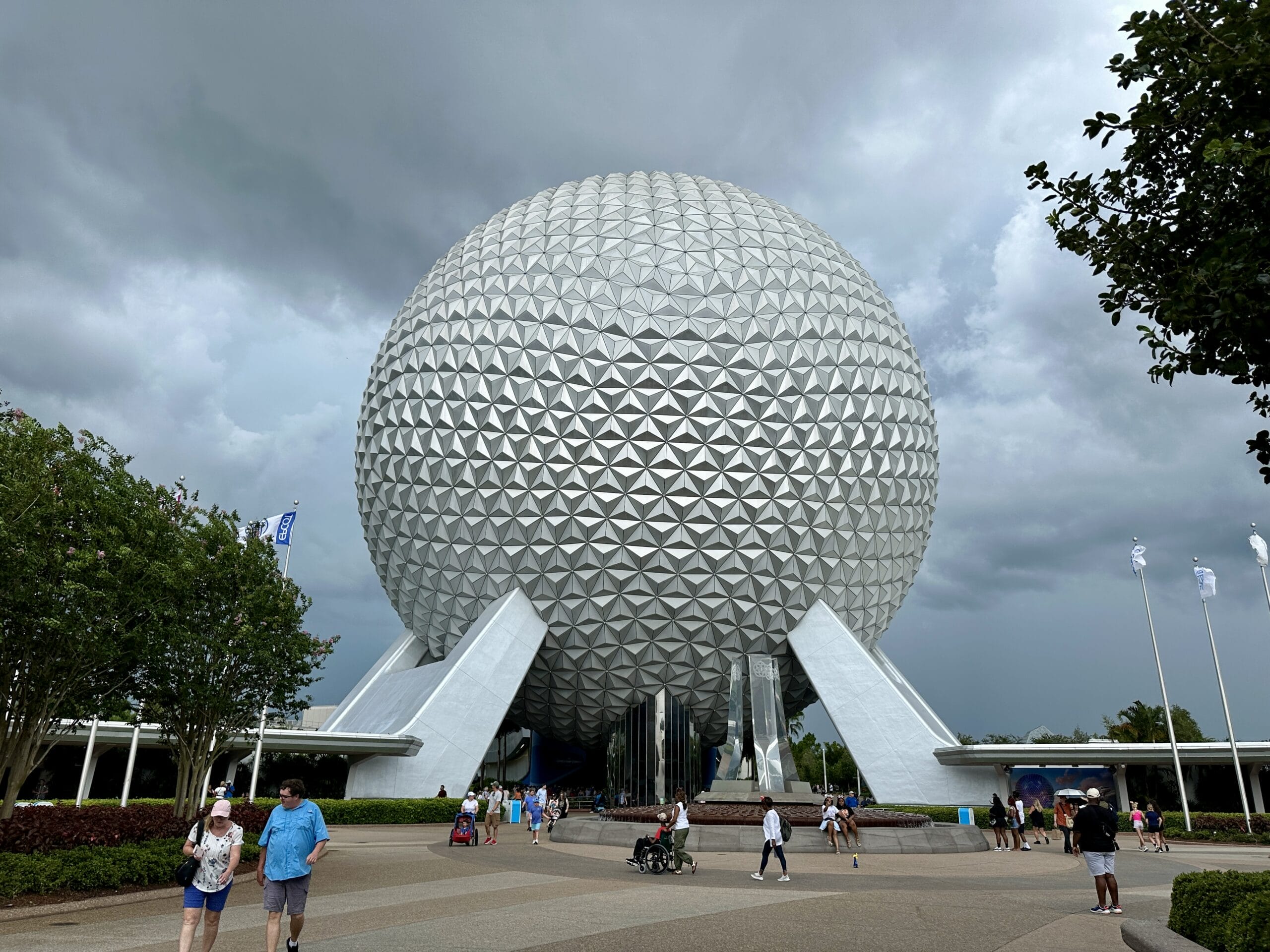 People walk near EPCOT’s iconic geodesic sphere, Spaceship Earth, with World Showcase visible under a cloudy sky.