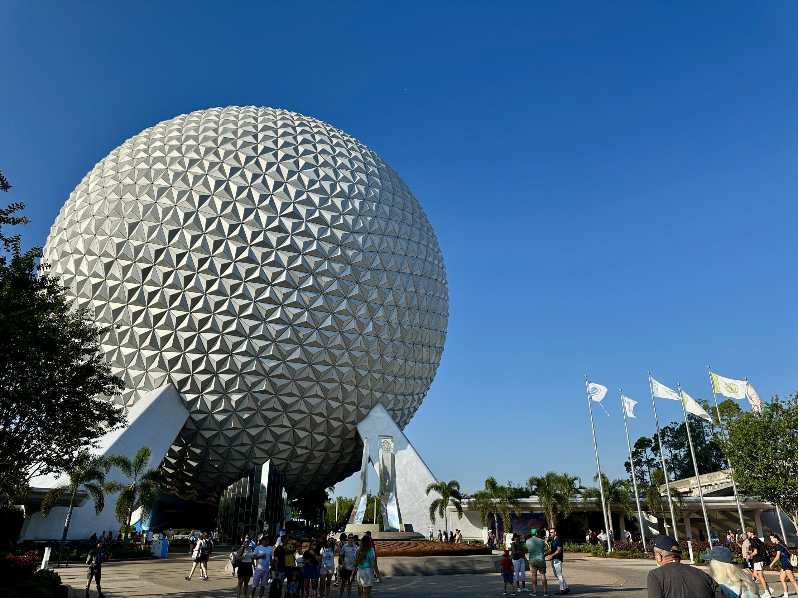 EPCOT’s Spaceship Earth geodesic sphere with visitors and flags near the World Showcase under a clear blue sky.