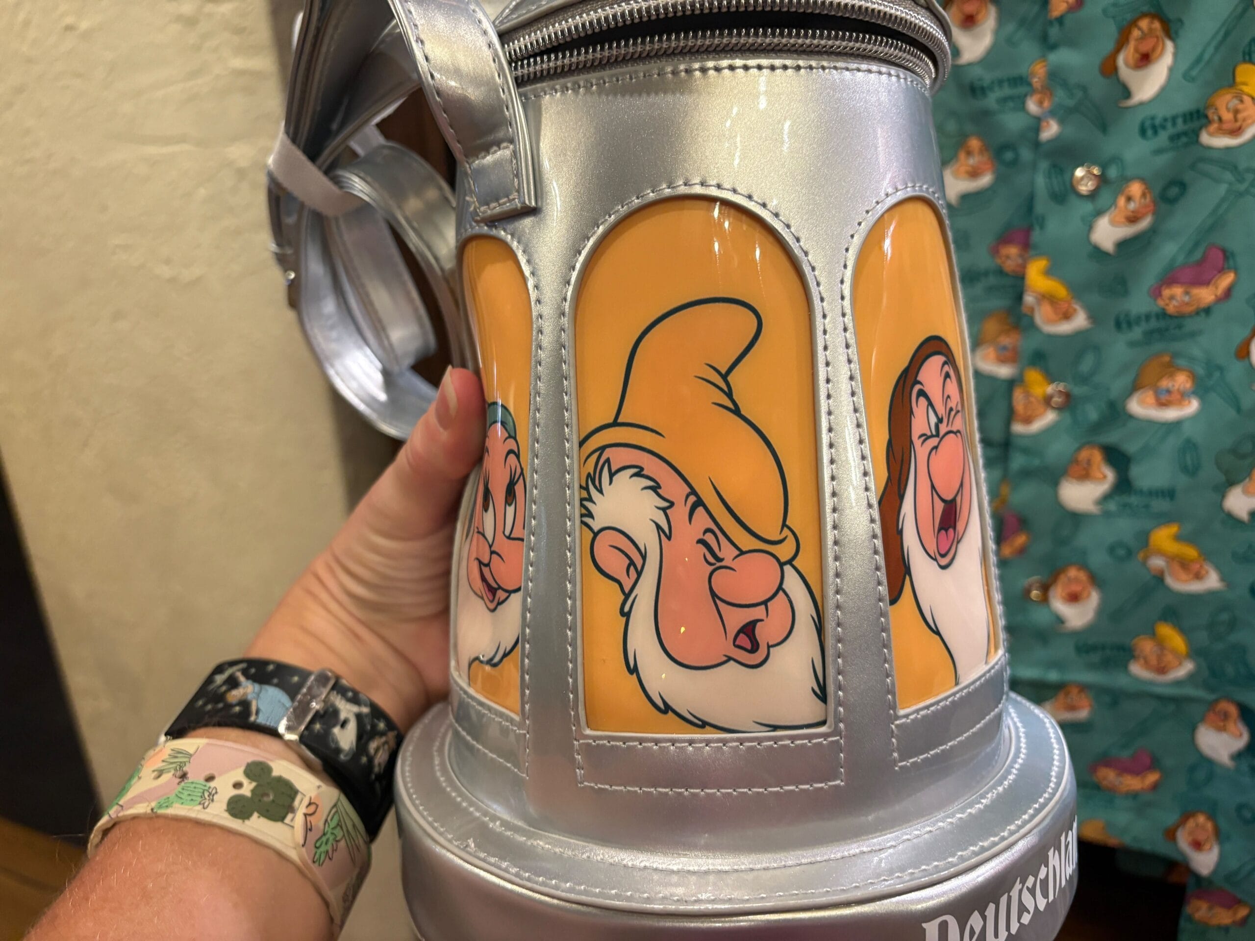 A hand holds a silver Stein Bag mug featuring EPCOT’s Seven Dwarfs, Grumpy and Happy, from Snow White.