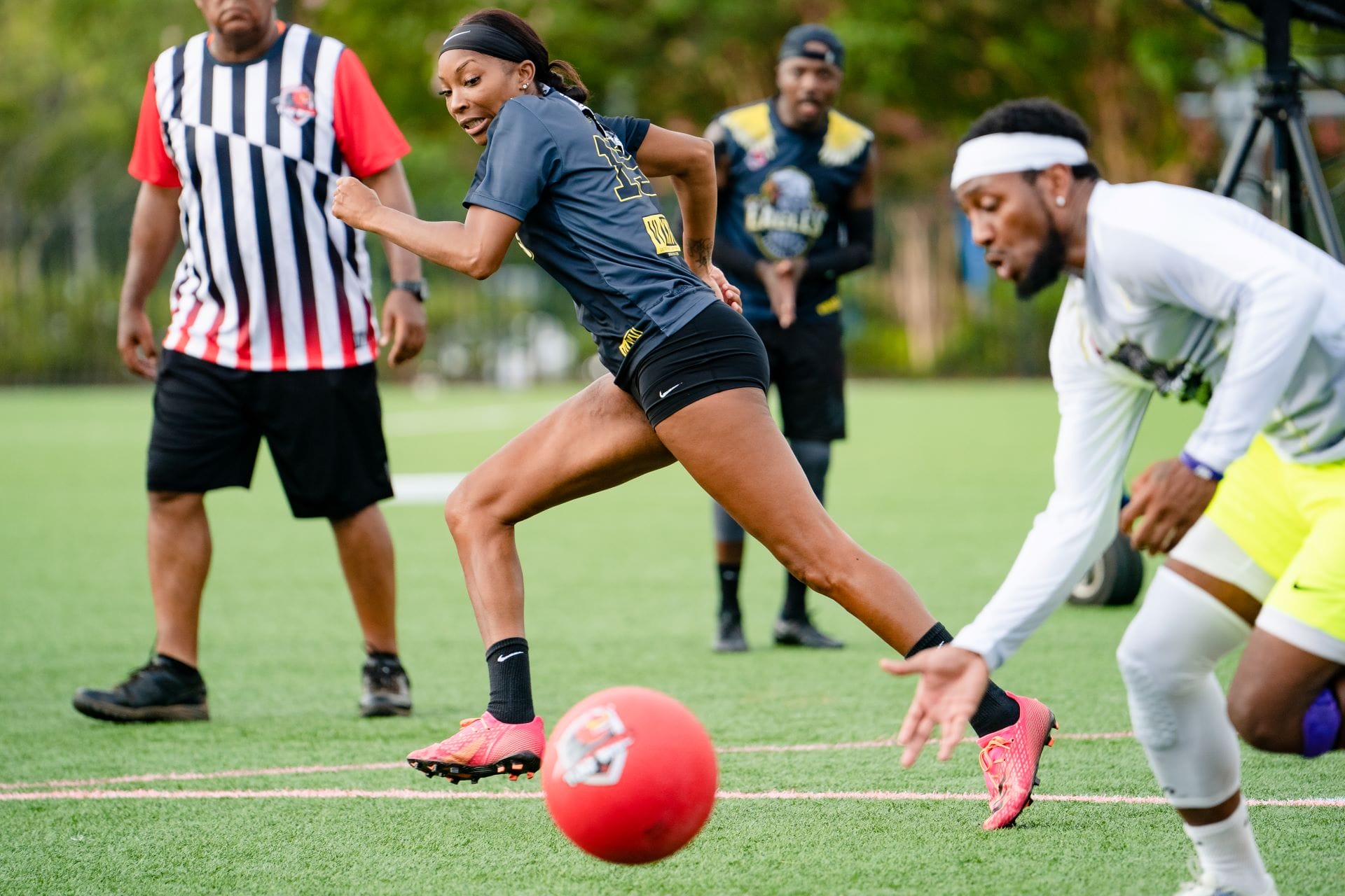 A woman runs toward a red kickball as other players chase in this lively game inspired by dodgeball on a grassy field.