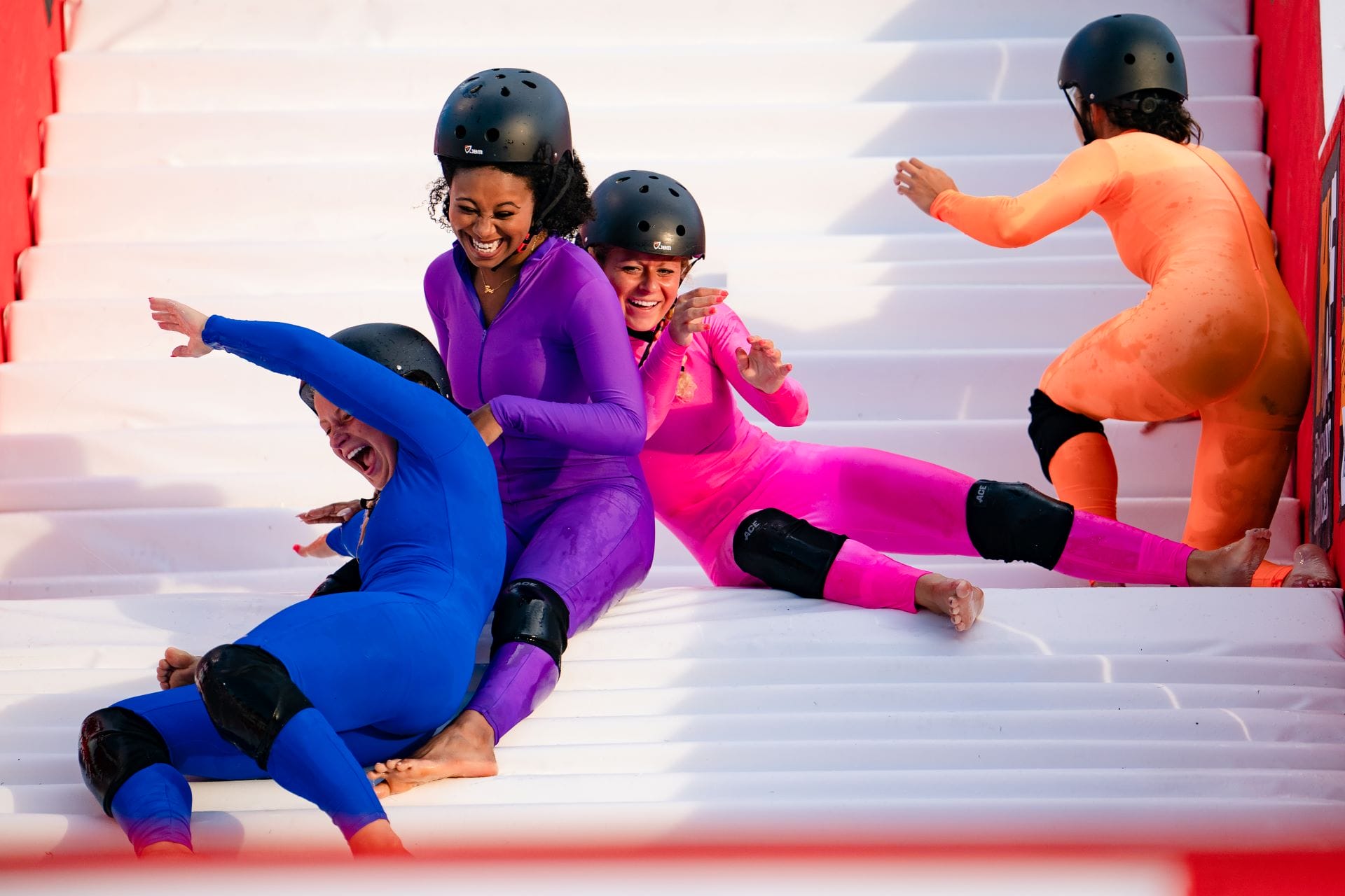 Four people in colorful suits and helmets laugh together on a Walt Disney World inflatable slide.