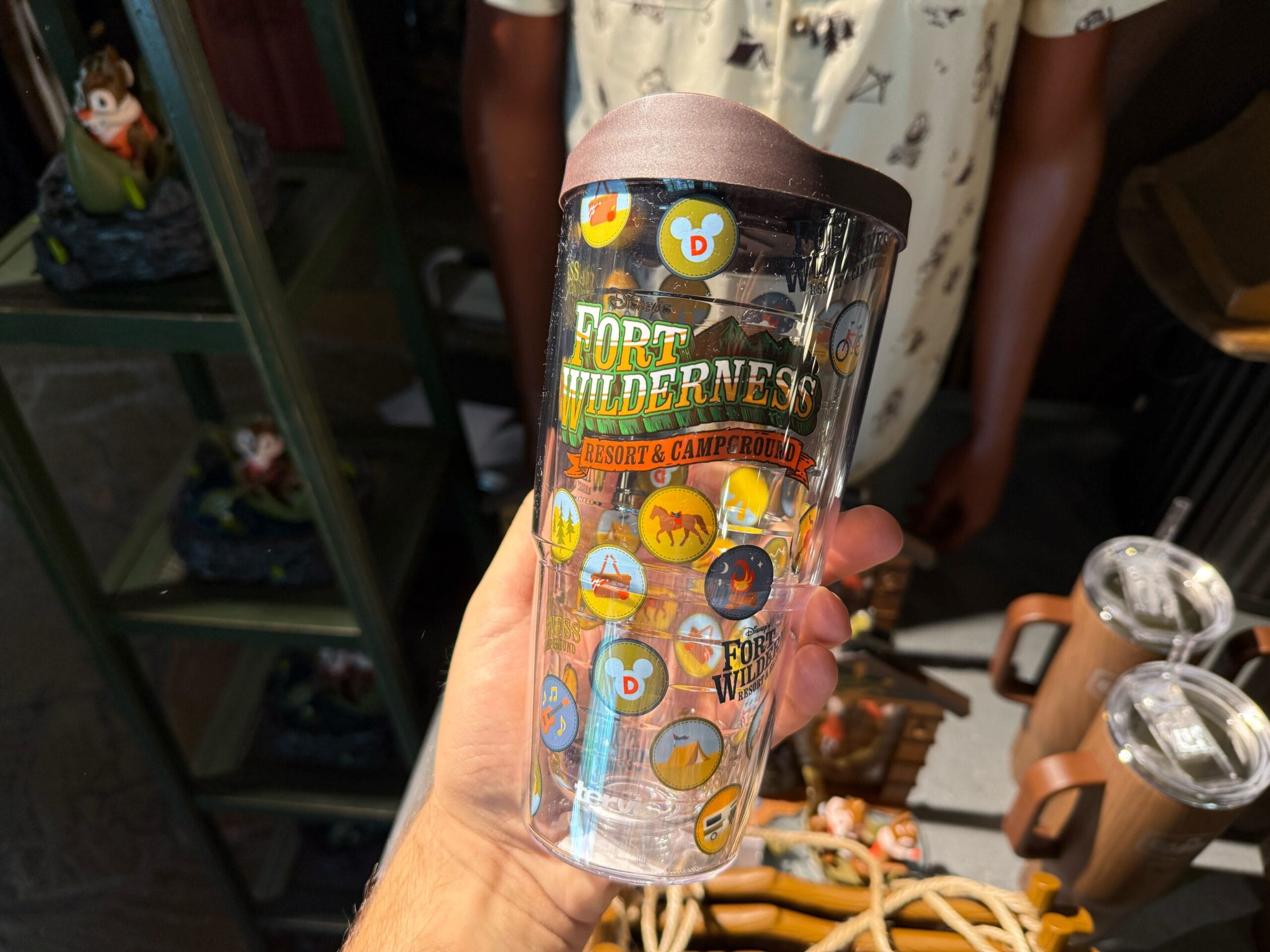 A hand holds Disney’s Fort Wilderness merchandise: a resort travel tumbler with colorful camp-themed icons and animals.