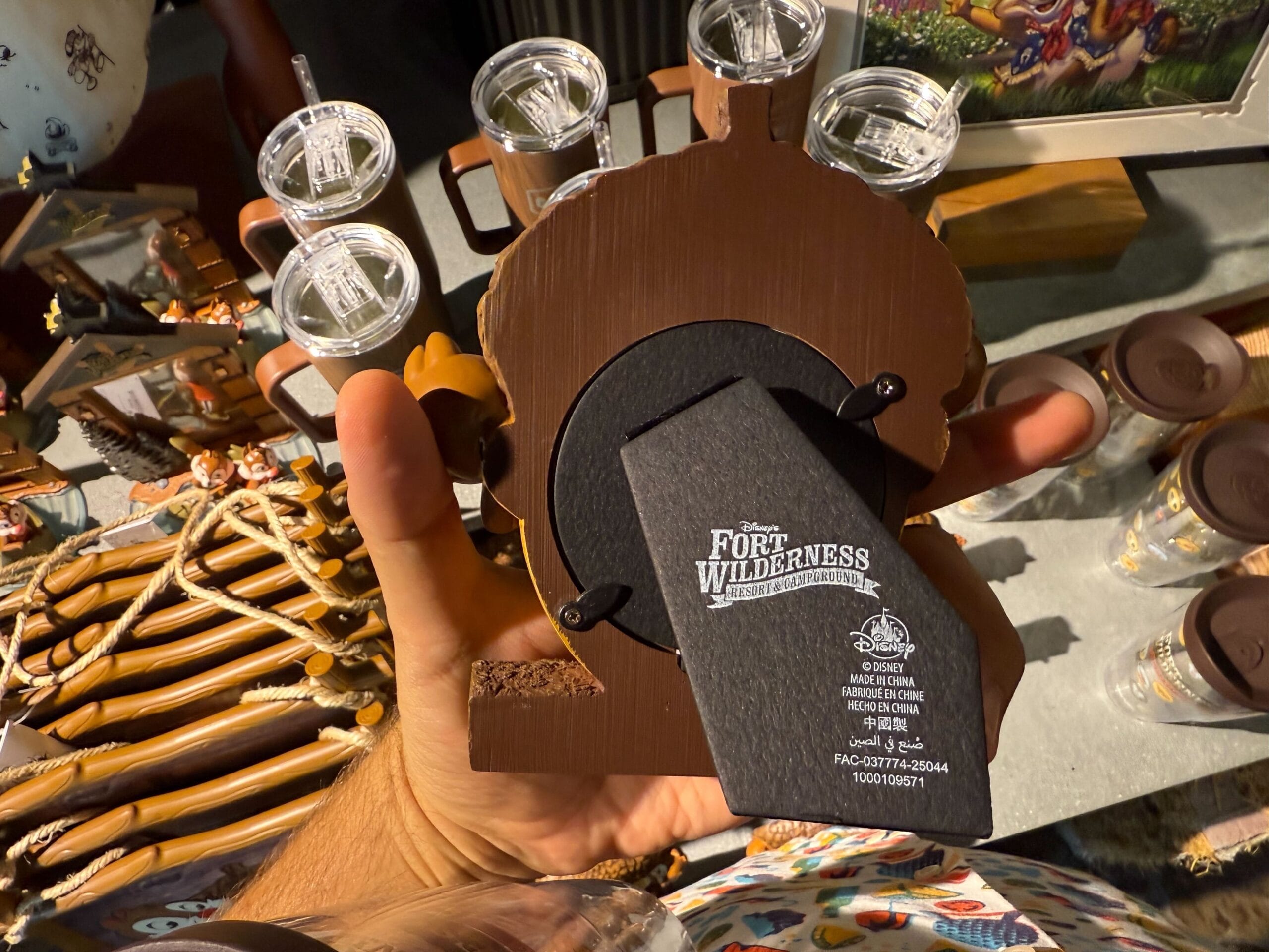 A hand holds Disney’s Fort Wilderness merchandise—a bear-shaped photo frame—with shot glasses in the resort background.