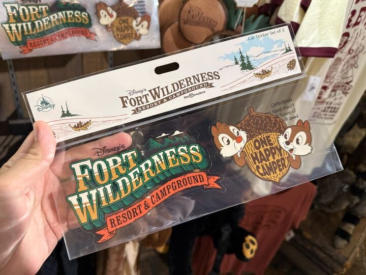 A hand holds Disney’s Fort Wilderness Resort merchandise—a car sticker set with Chip and Dale and a happy camper acorn.