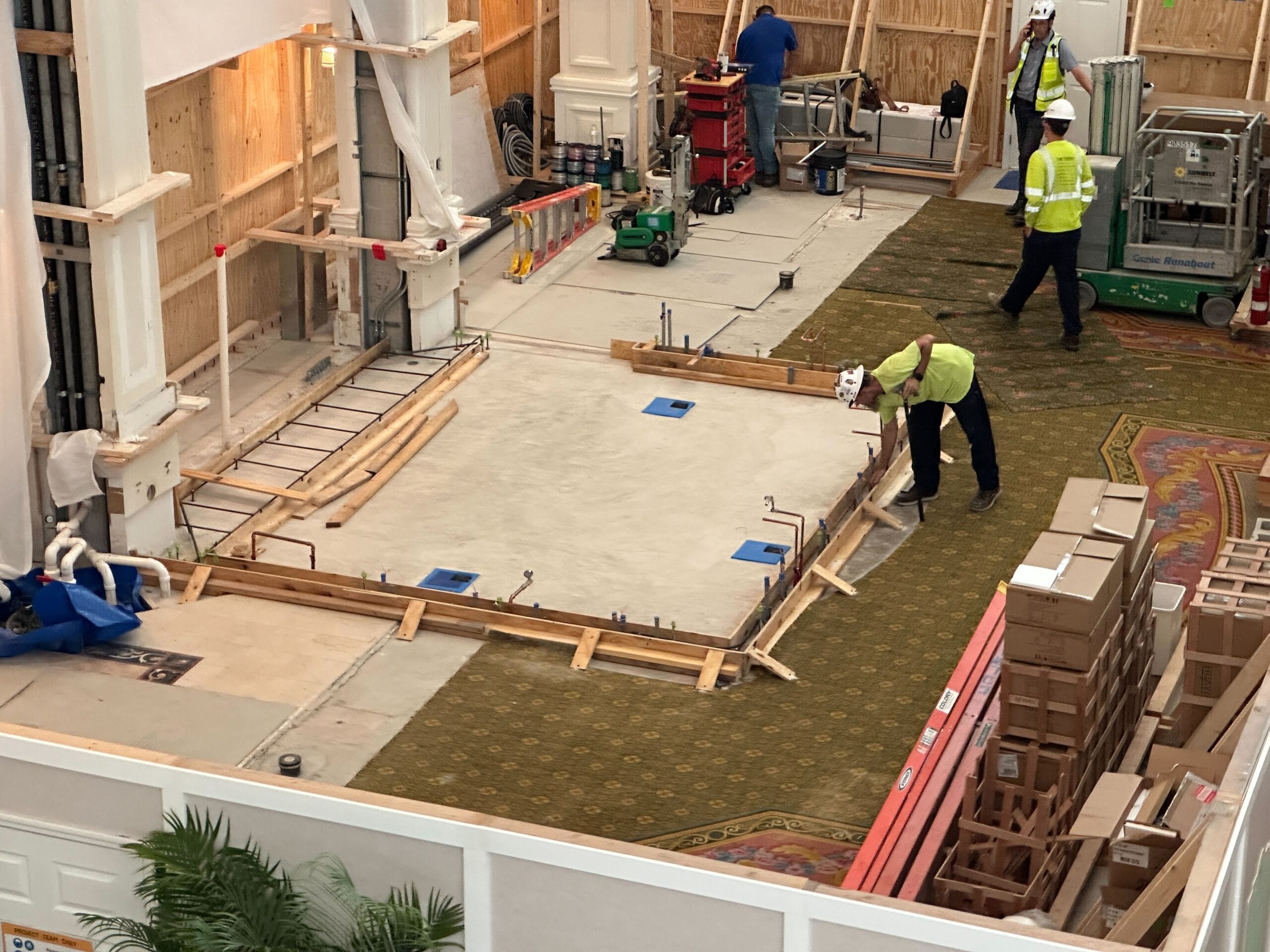 Workers in safety vests build and inspect a concrete platform during a Grand Floridian Resort update indoors.