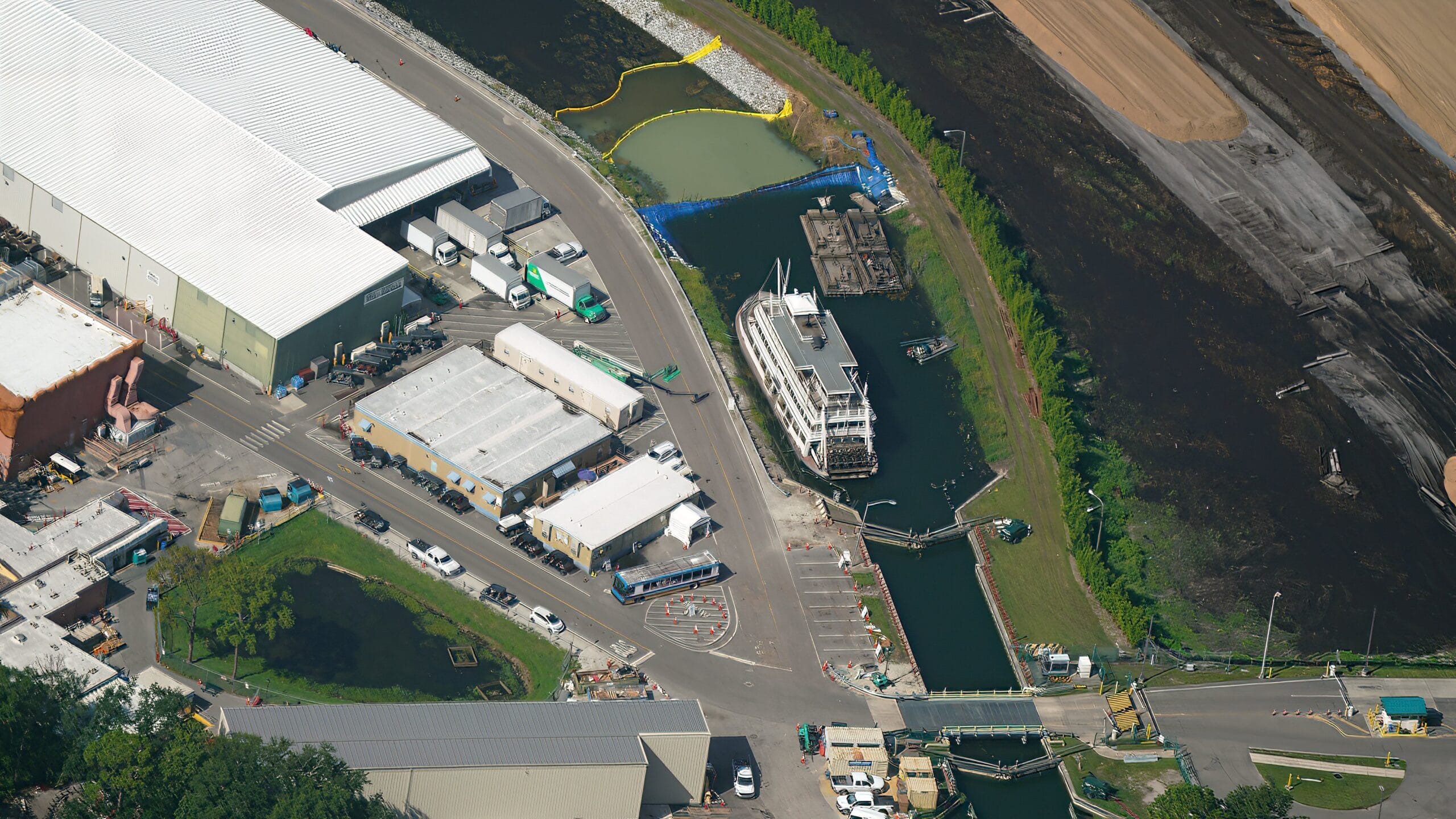 Aerial view of a Liberty Square Riverboat docked in a canal near industrial buildings and parked vehicles.