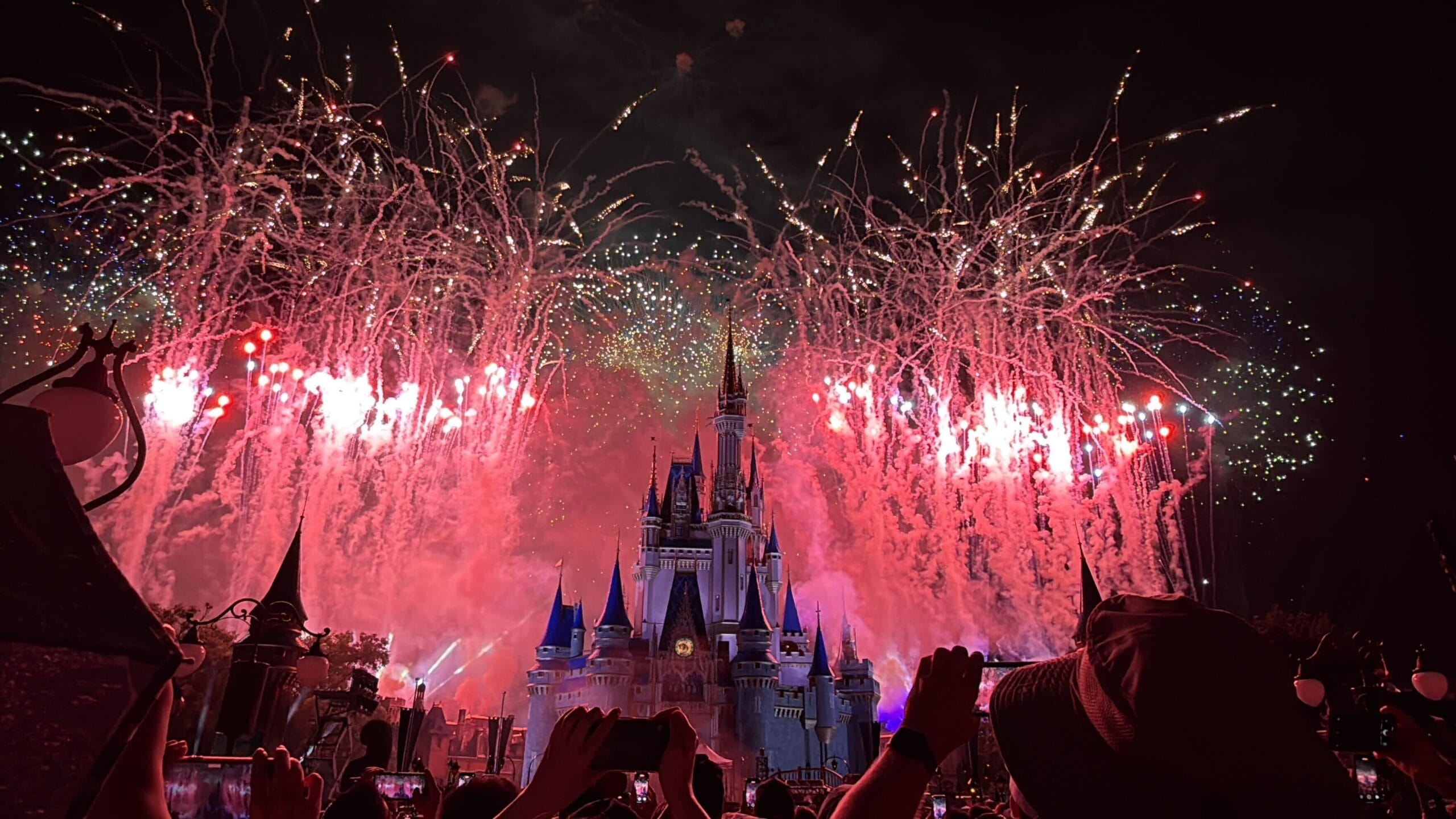 Colorful fireworks burst over a lit-up castle at night as people celebrate America and take photos in the foreground.