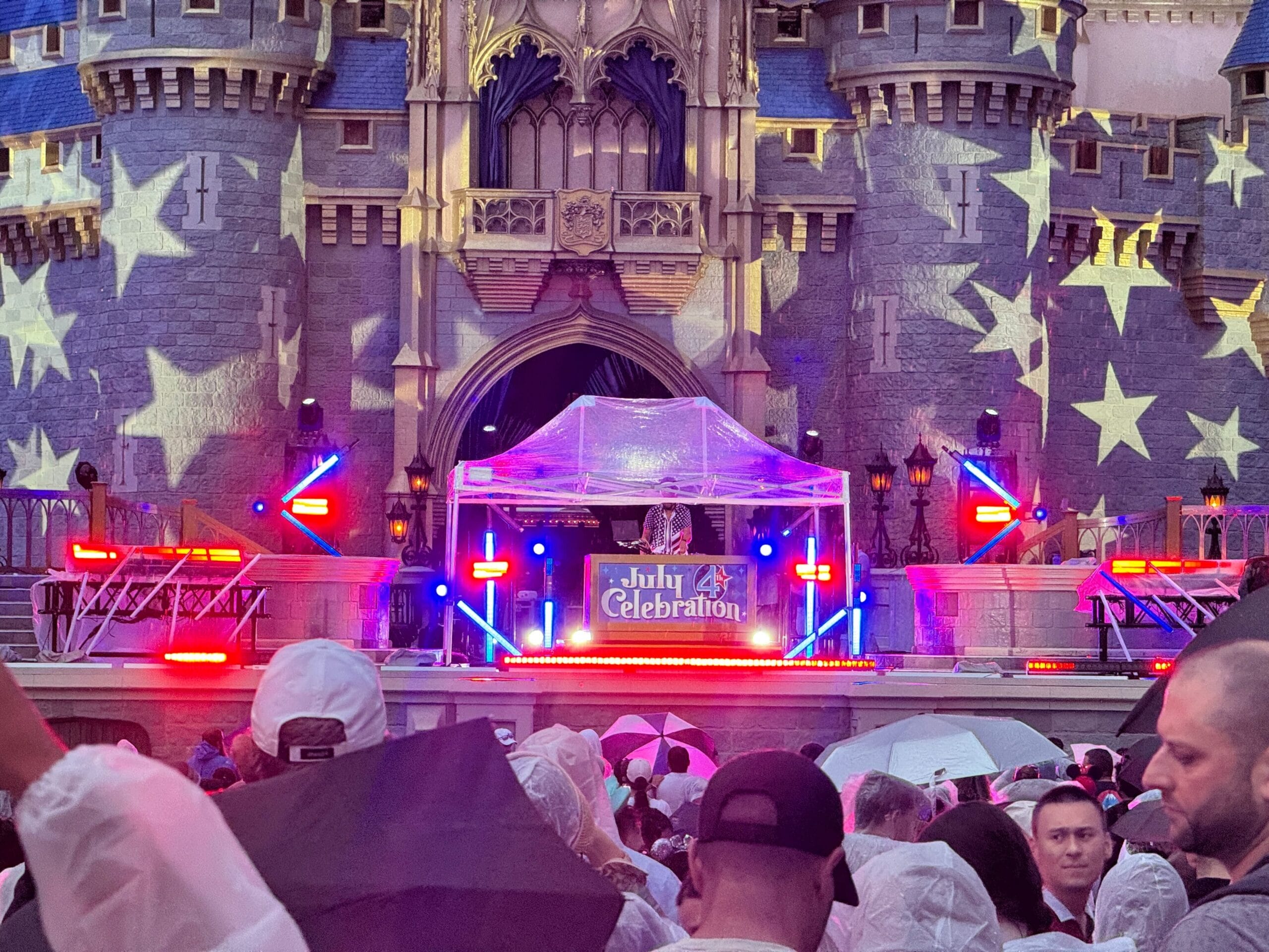 A DJ performs at a July 4th Celebration as fireworks light up the sky, with a crowd in rain ponchos watching.