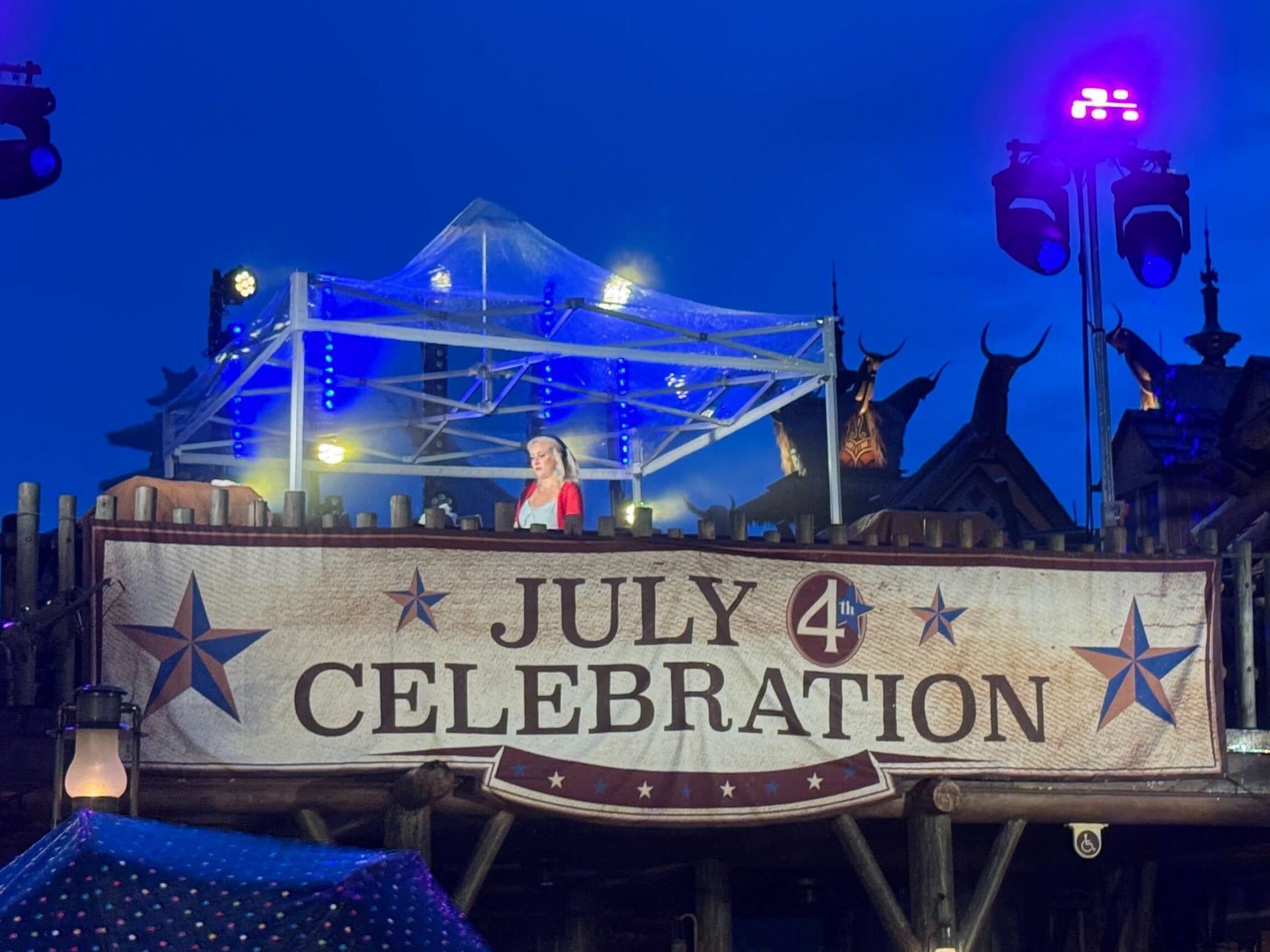 A woman stands on a balcony under lights behind a large "July 4th Celebration" sign as fireworks sparkle at dusk.