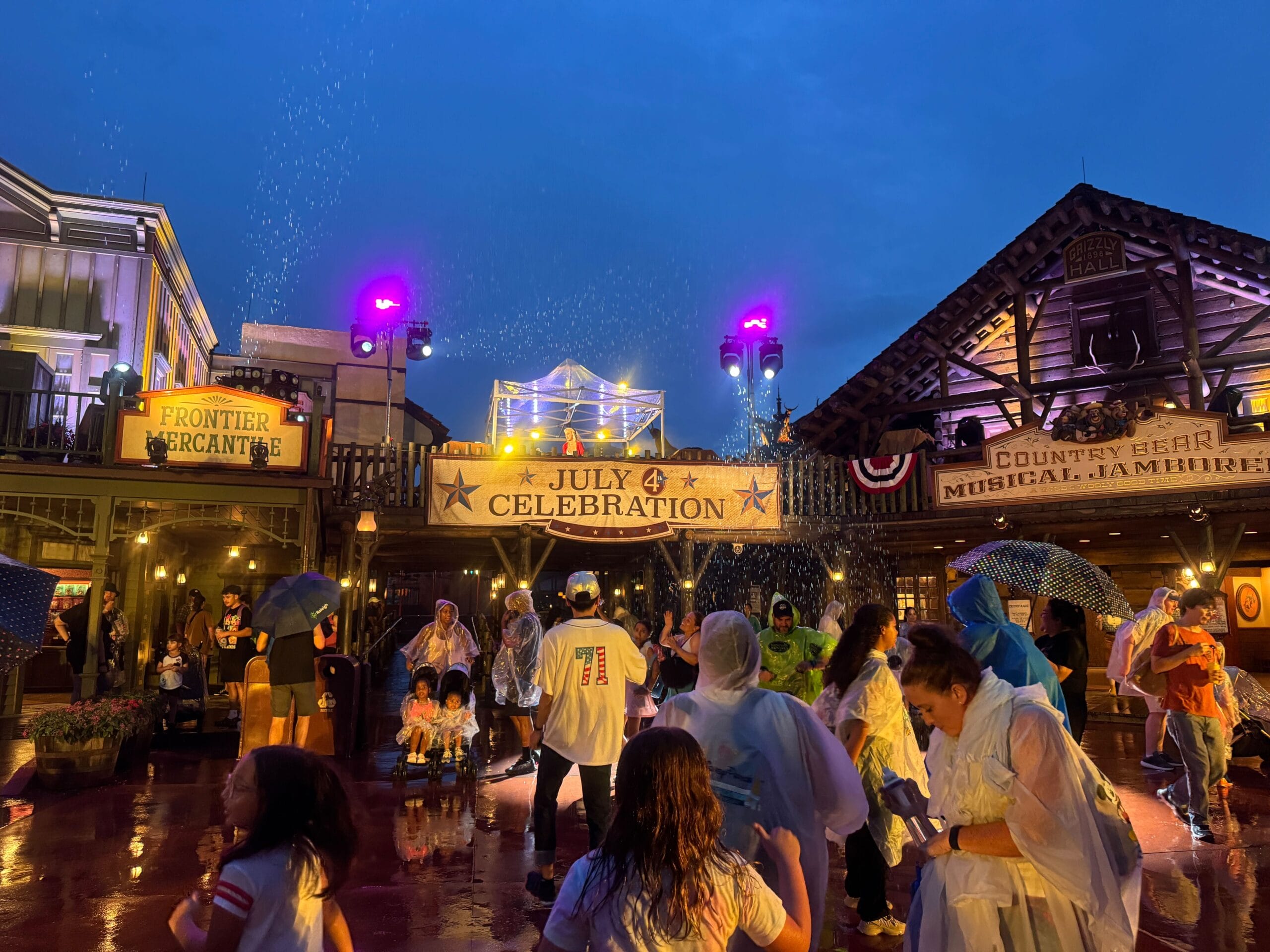 People in rain ponchos celebrate America under lights and water sprays at a lively, western-themed Fourth of July 2025 venue.
