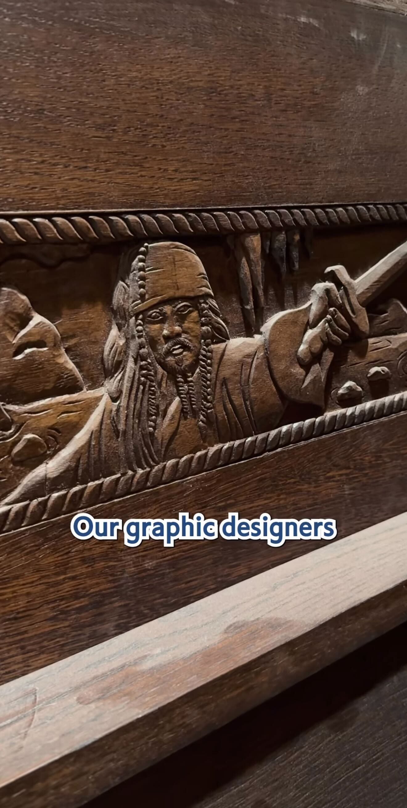 Wooden carving of a pirate at The Beak and Barrel with "Our graphic designers" in bold white text overlaying the image.