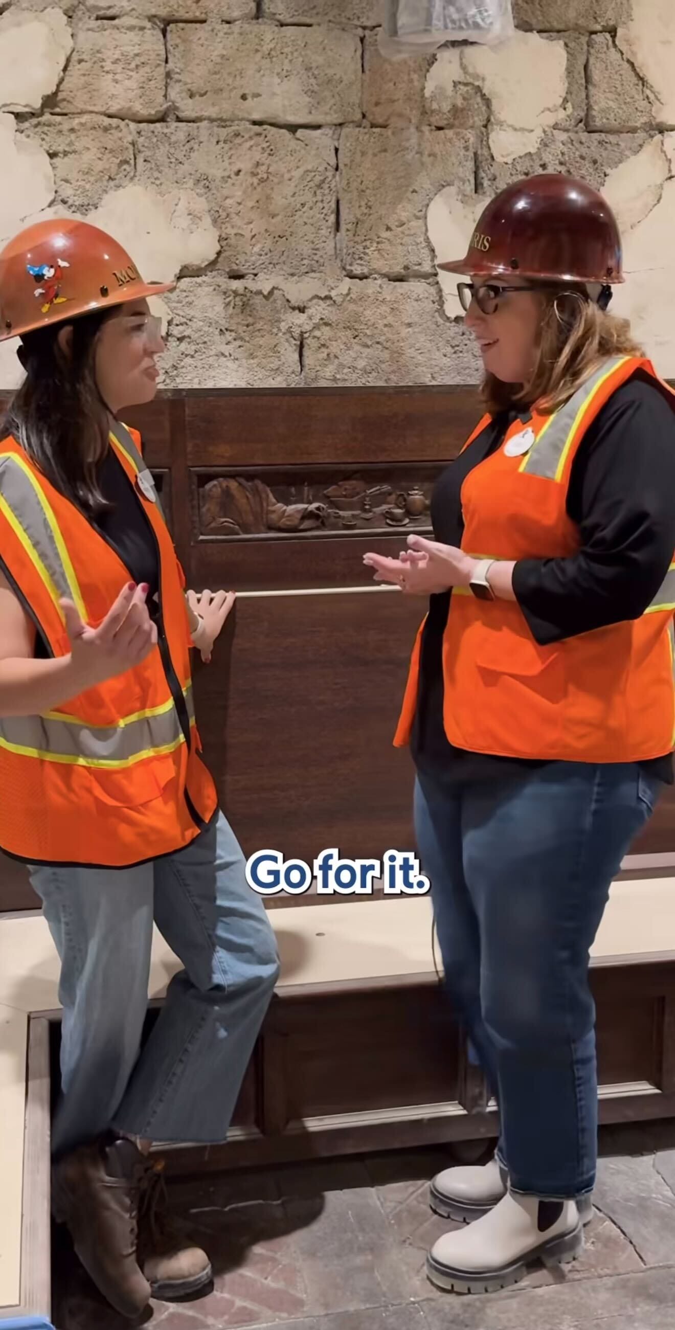 Two women in orange safety vests and hard hats talk by a stone wall. Text reads, “Go for it, Magic Kingdom spirit.”