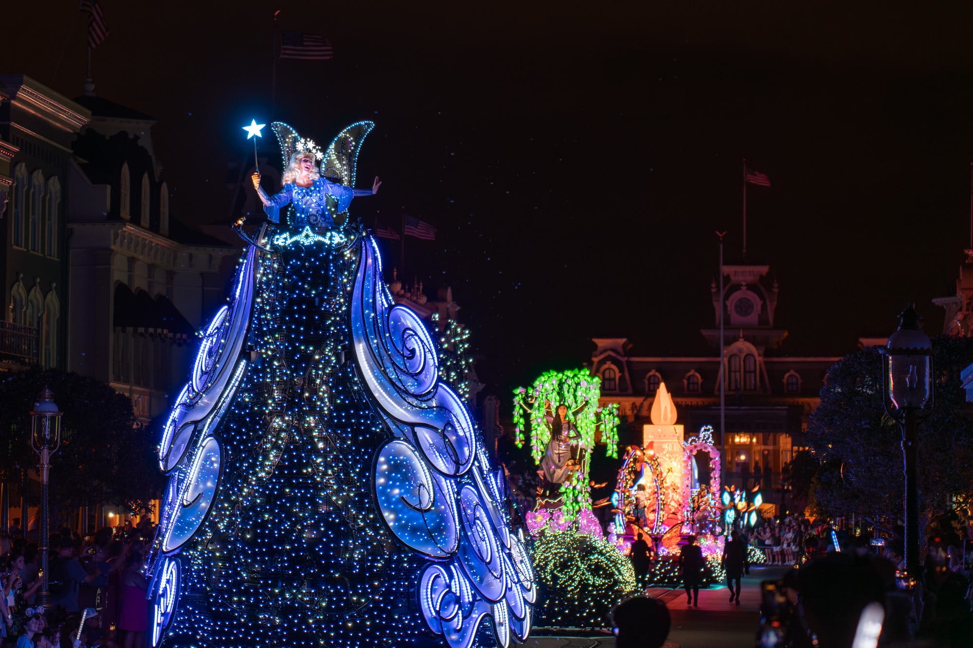 A performer in a glowing blue fairy costume rides a float during Disney Starlight Parade performances at night.