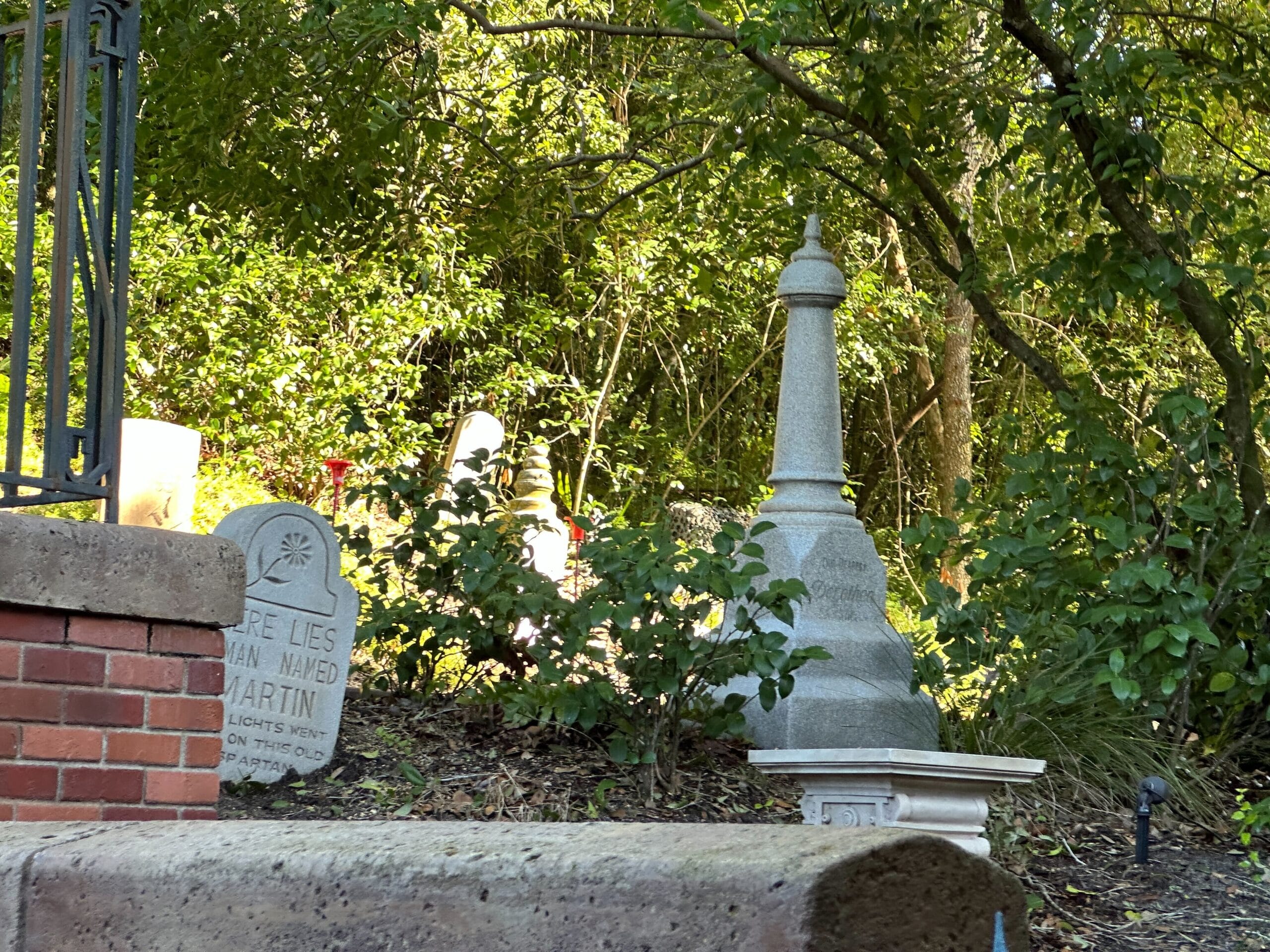 Several gravestones and monuments stand among green trees, like a scene from Magic Kingdom’s Haunted Mansion.