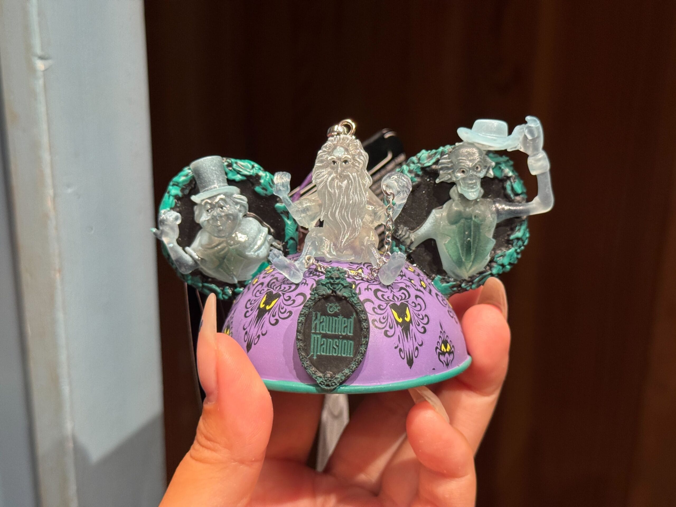 Hand holding a Haunted Mansion-themed Mickey ears ornament, a must-have Magic Kingdom merch with ghostly figures and vibrant details.