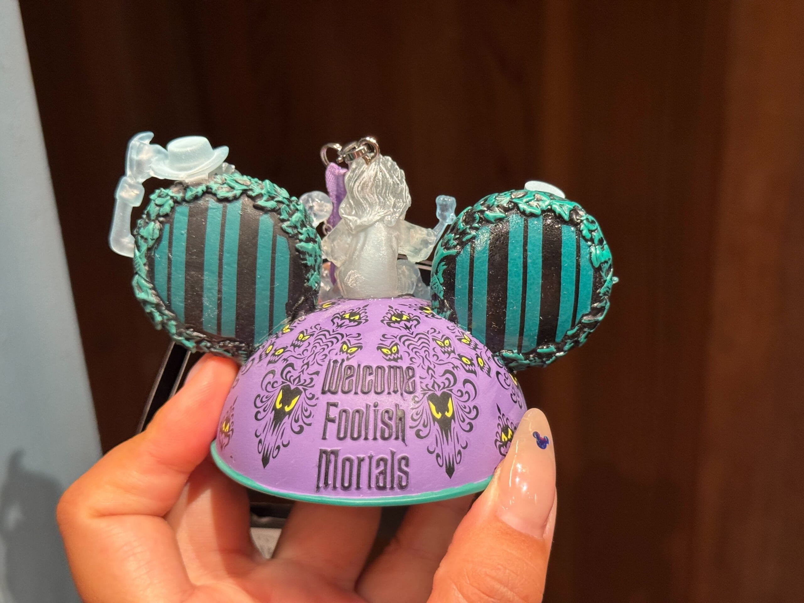 Hand holding Haunted Mansion-themed merch: Mickey ears hat reads “Welcome Foolish Mortals” at Magic Kingdom.