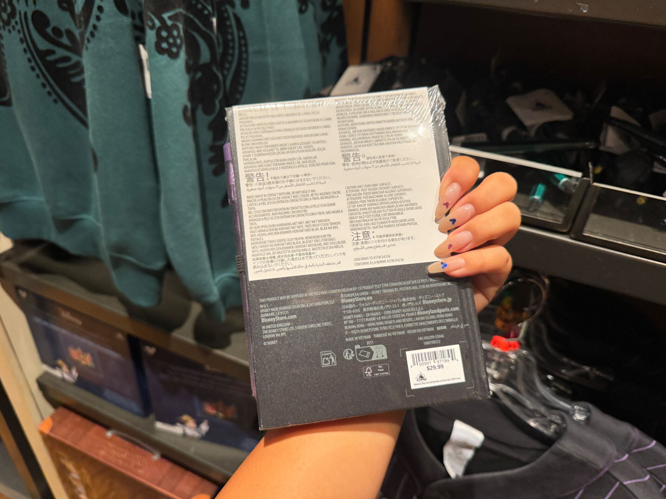 A hand with painted nails holds Haunted Mansion merch with multilingual instructions in a Magic Kingdom clothing store.