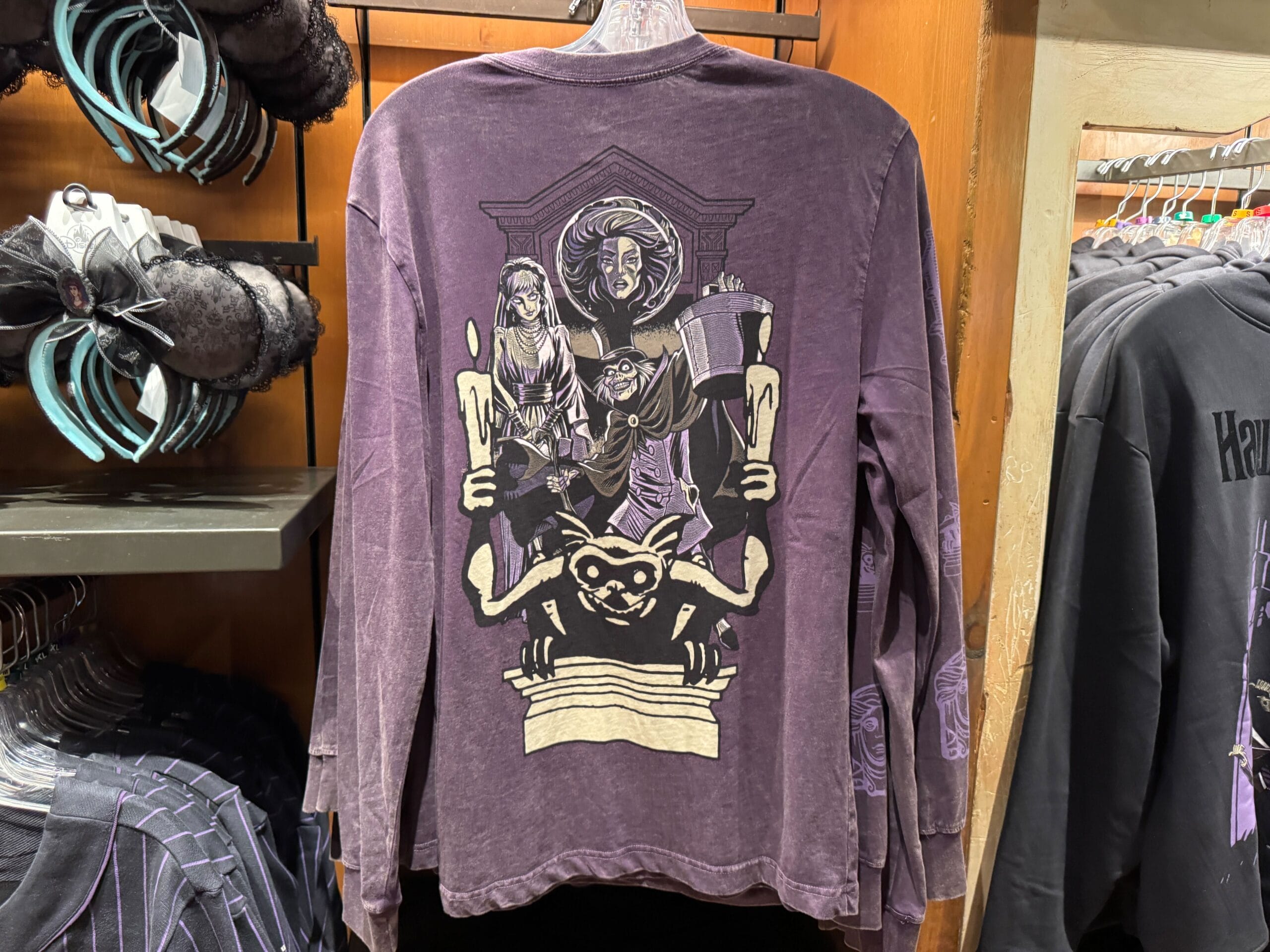 A purple long-sleeve Merch shirt features gothic Haunted Mansion artwork with women, candles, and a gargoyle on the back.
