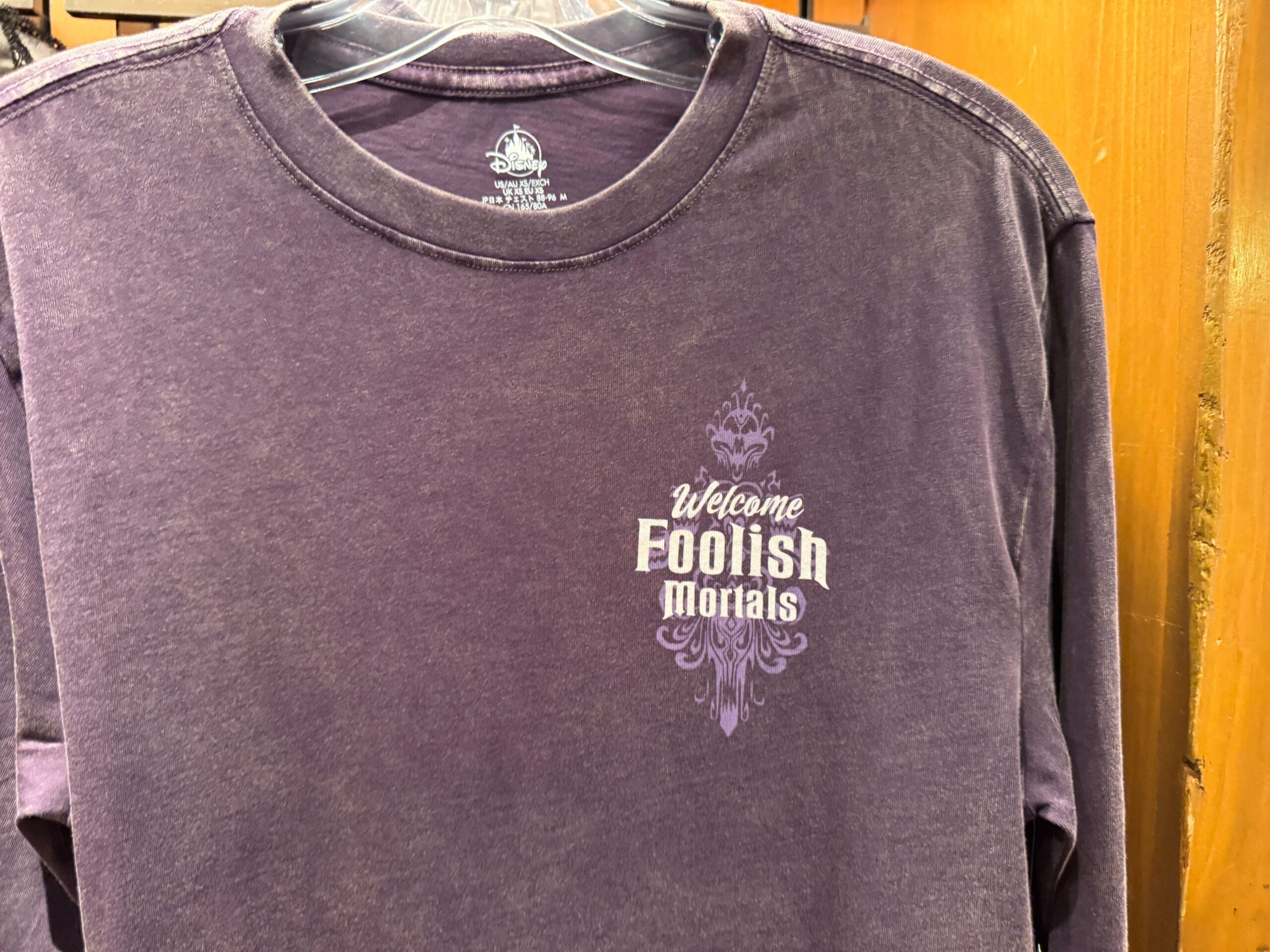 A purple long-sleeve Haunted Mansion shirt with "Welcome Foolish Mortals" in white decorative font on the chest.