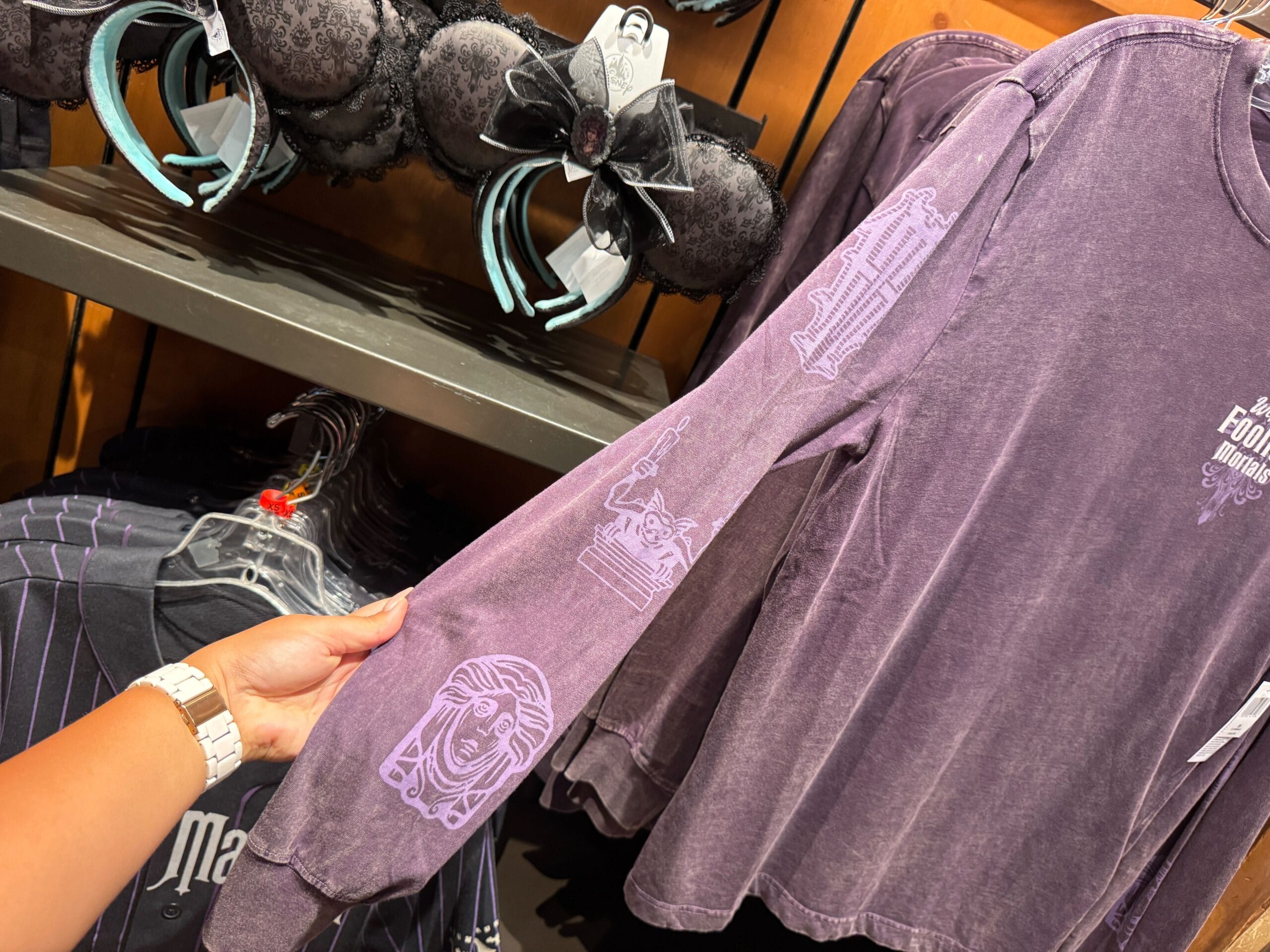 A hand holds a Haunted Mansion-themed purple long-sleeve shirt in a Magic Kingdom merch store.