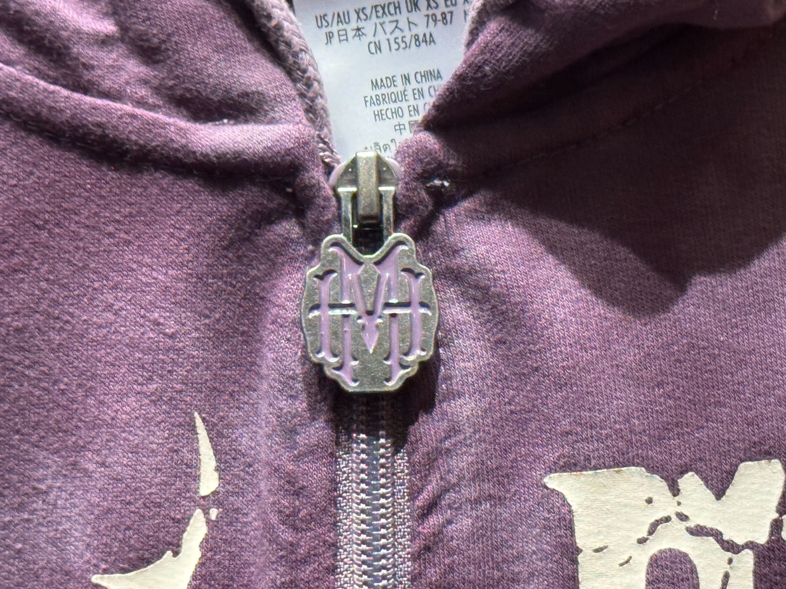 Close-up of a hoodie zipper with a decorative "M" pull, inspired by Haunted Mansion merch on purple fabric.