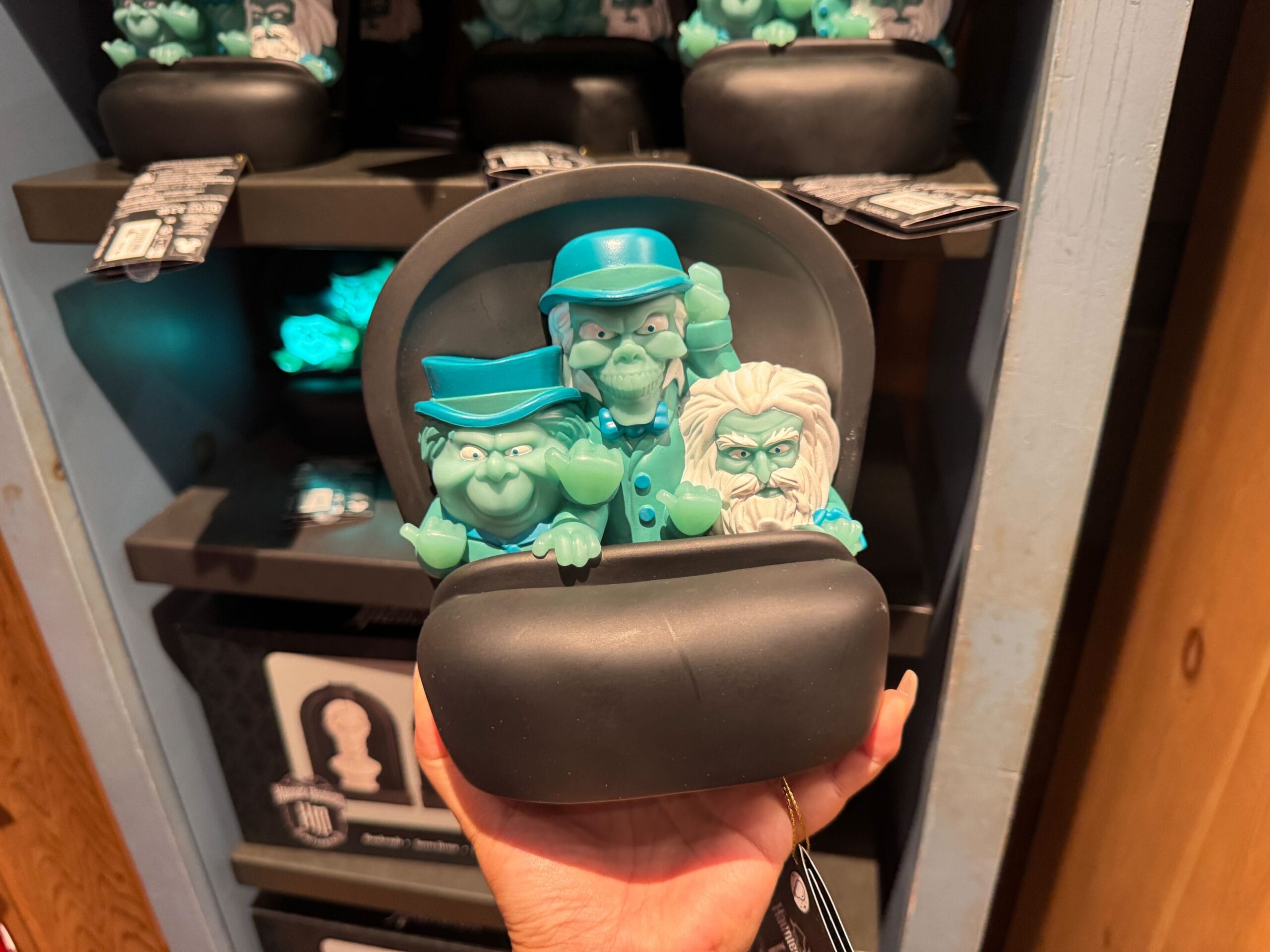 A hand holds Haunted Mansion merch—a figurine of three green ghosts in a black doom buggy from Magic Kingdom.