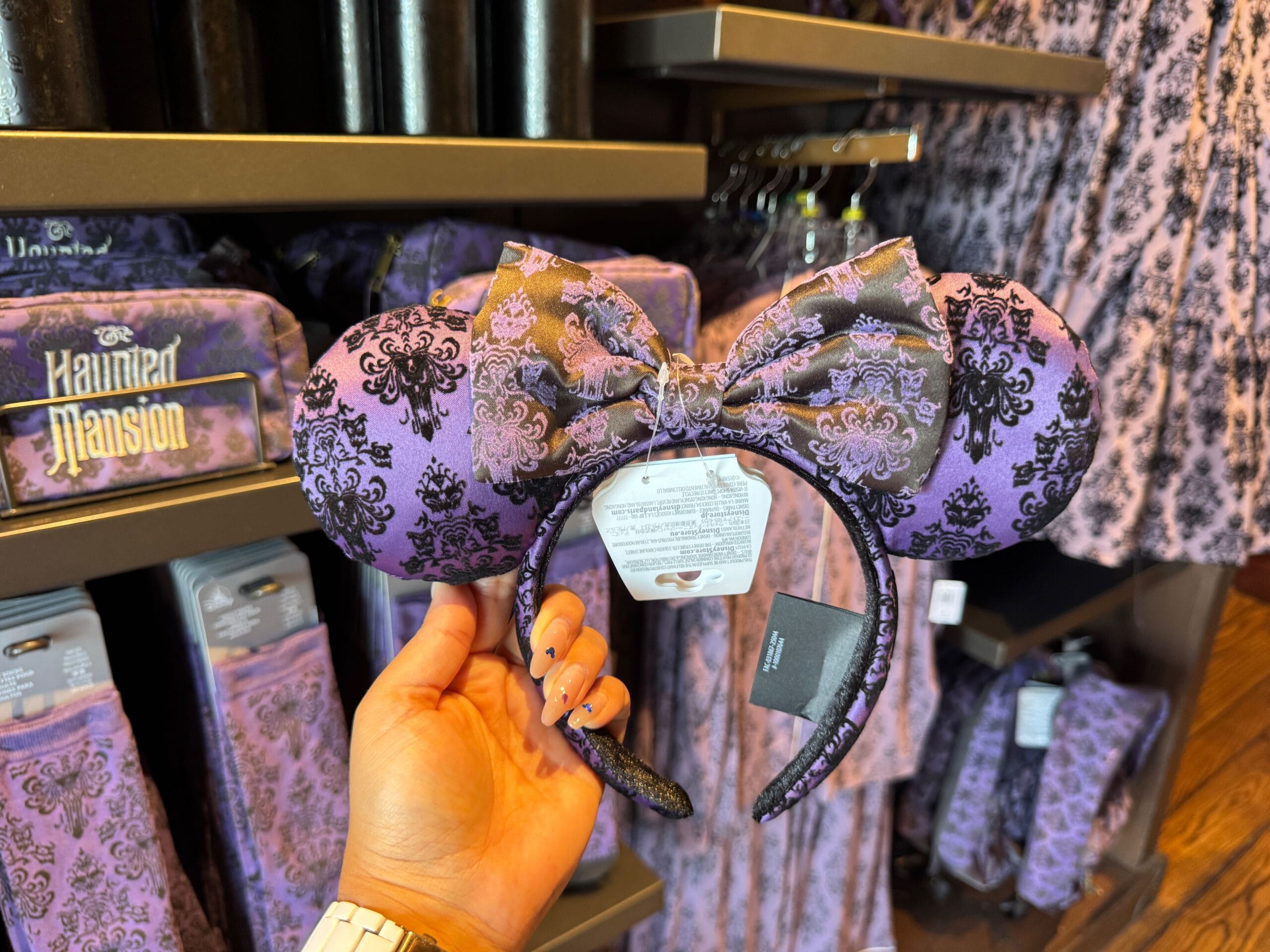 A hand holds a purple Haunted Mansion Minnie Mouse ear headband amid matching merch at Magic Kingdom.